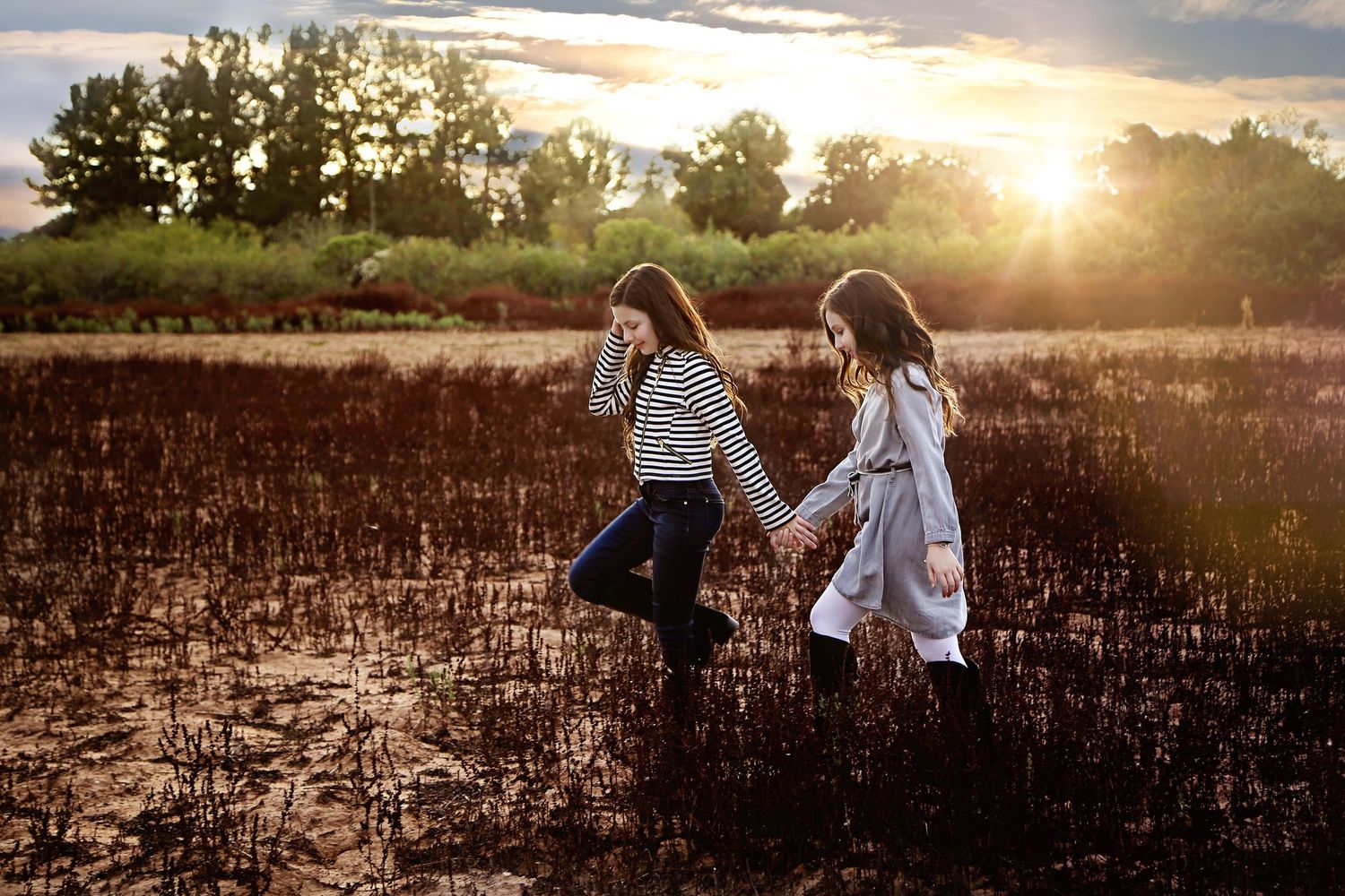 Two girls hold hands and walk across a charred field at golden sunset with trees glowing in the background.