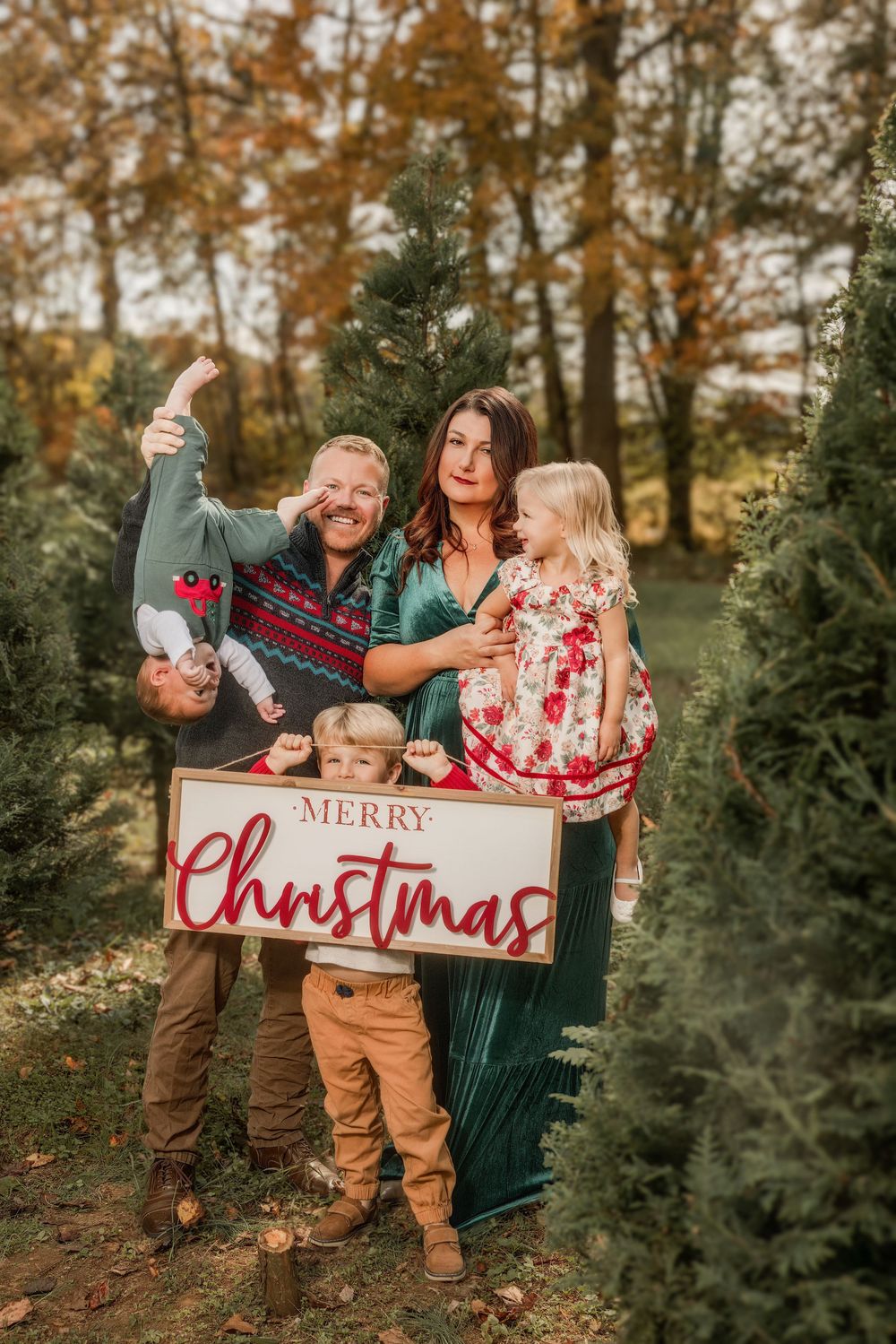 Family of four posing with a Merry Christmas sign among evergreen trees during a festive holiday photo session.