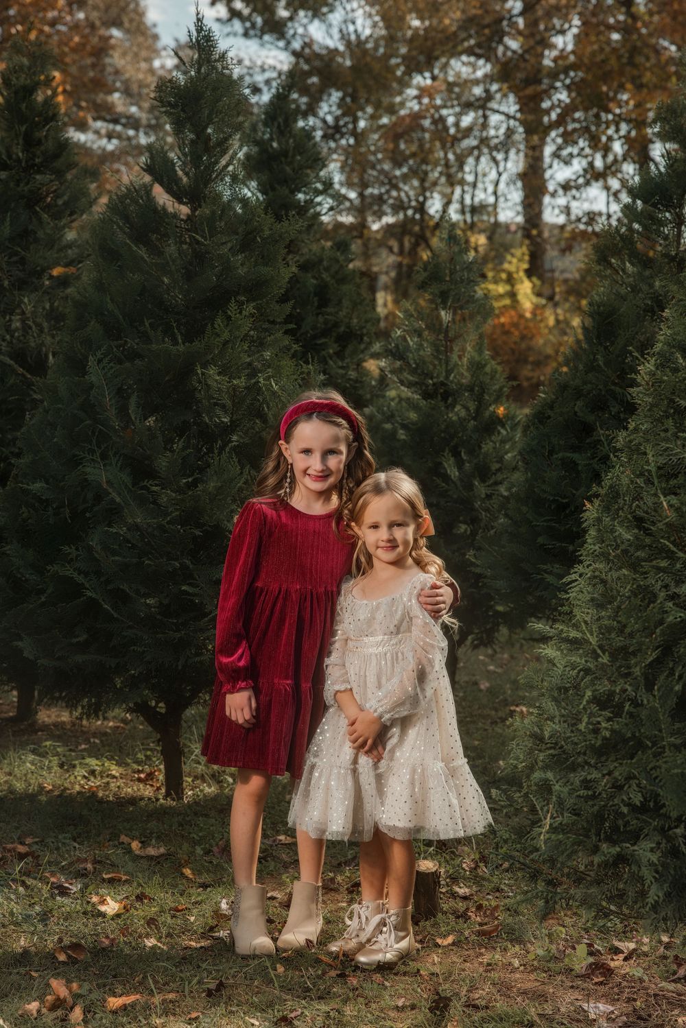 Two young girls in festive dresses pose among Christmas trees at a tree farm in autumn.