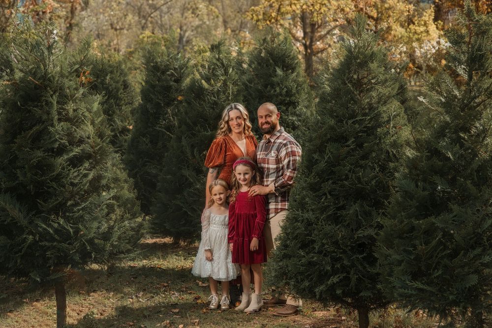 Family of four posing among Christmas trees at a tree farm, parents with two young daughters in festive holiday outfits.