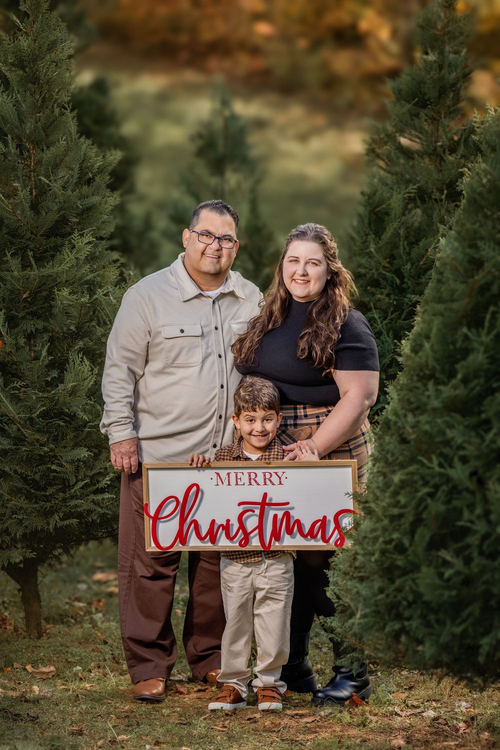 Family of three posing with a Merry Christmas sign at a Christmas tree farm during a festive holiday photo session.