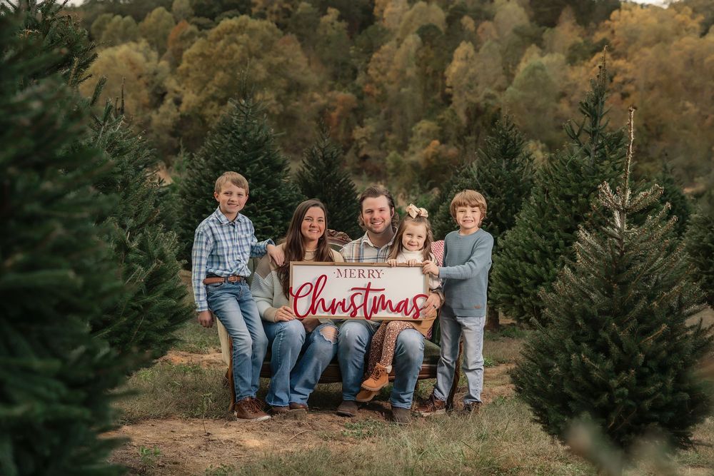 Family of five poses with Merry Christmas sign at a Christmas tree farm for a festive holiday photo.