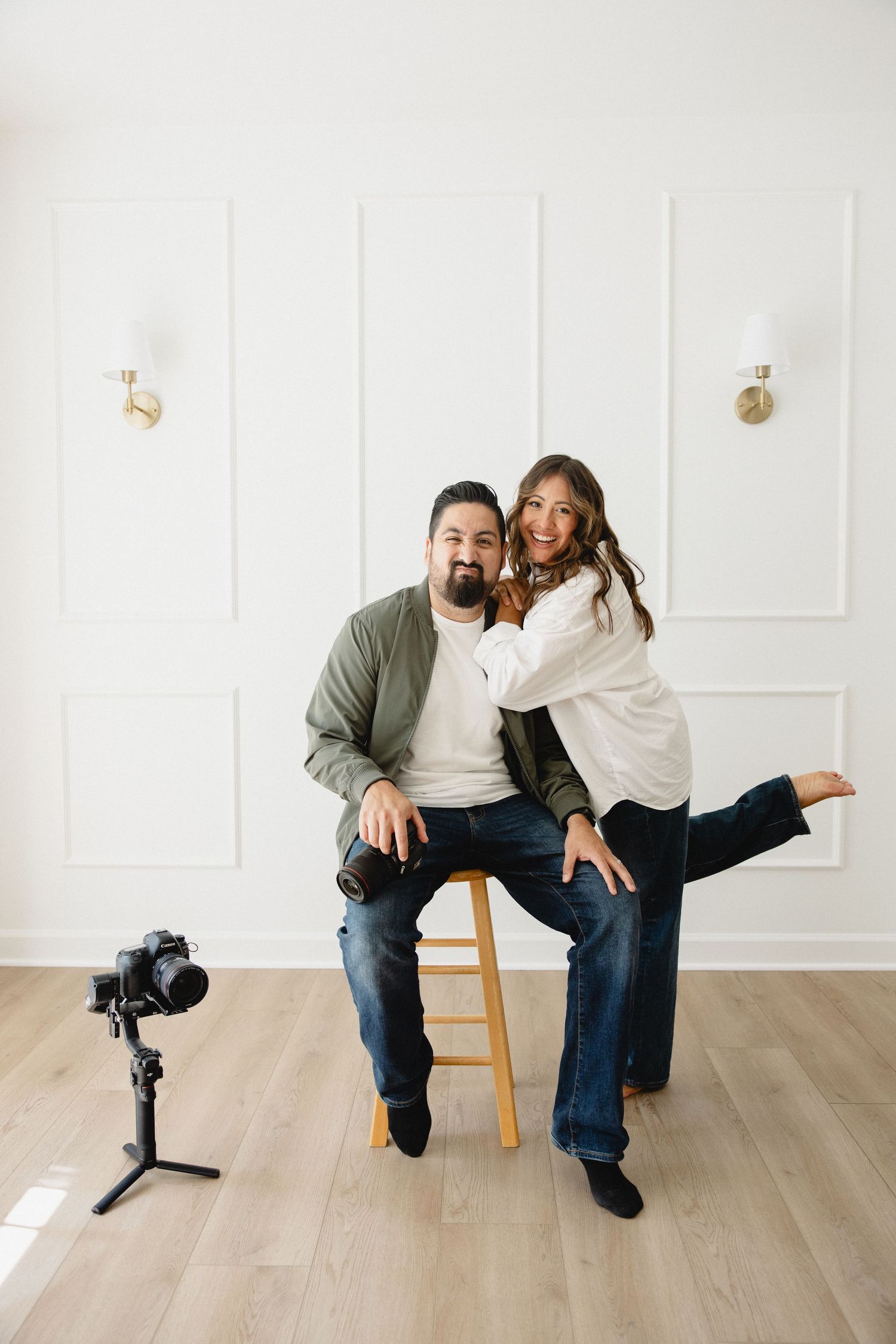 A smiling couple poses in a bright studio, man seated on stool, woman leaning beside him, camera on tripod nearby.