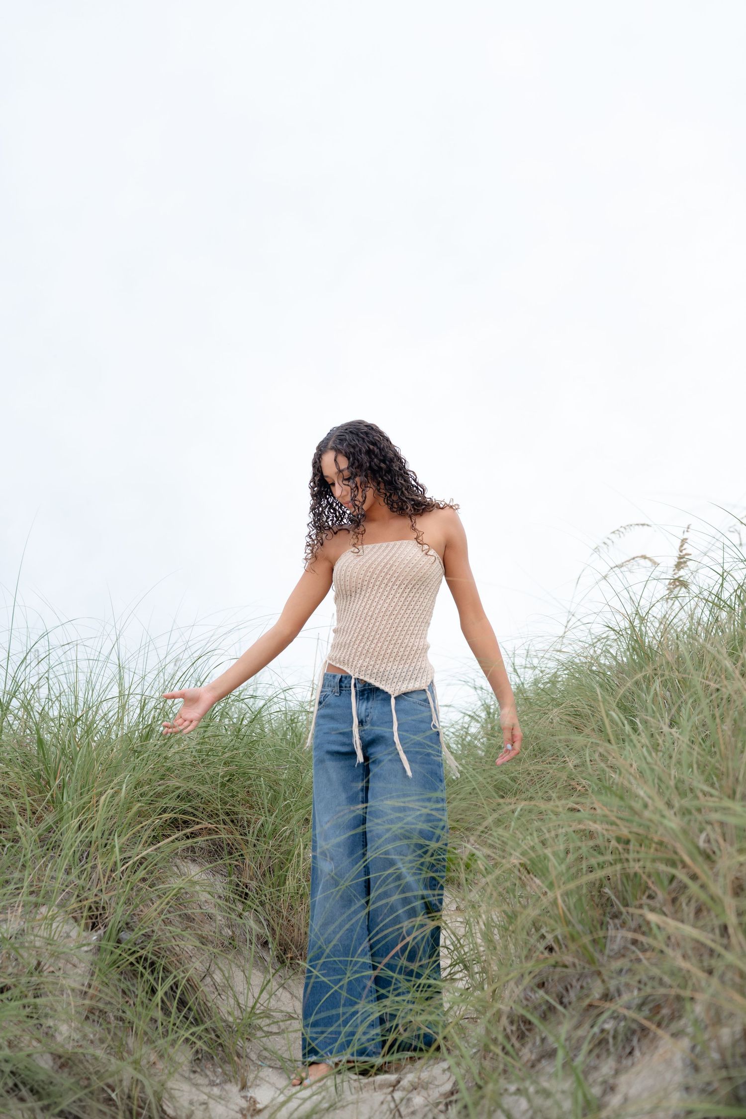 Young woman in knit tube top and wide-leg jeans standing among beach grass on sandy dunes on overcast day.