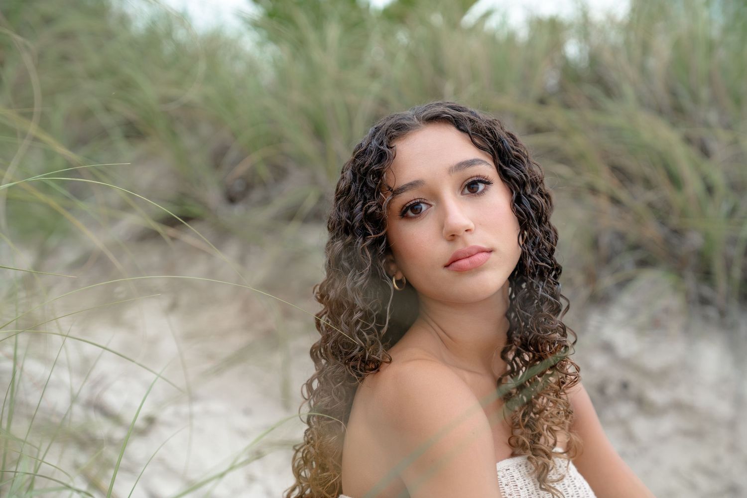 Young woman with curly hair poses among beach dunes and tall grass in a serene outdoor portrait.