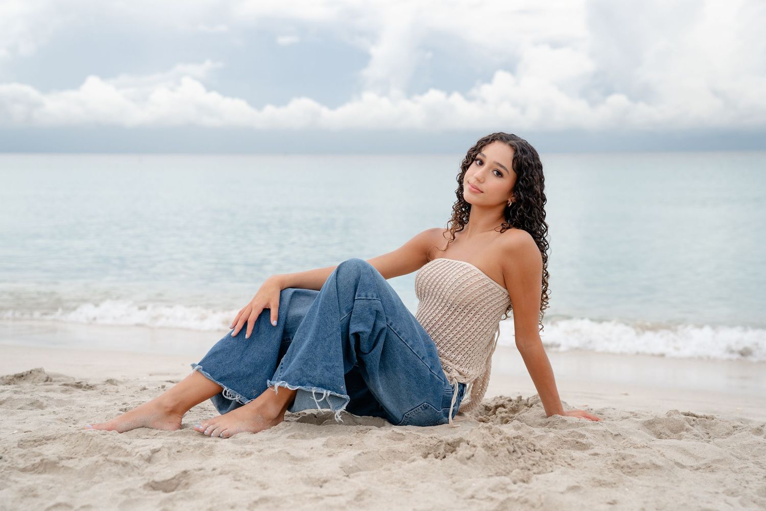 Young woman with curly hair sits on sandy beach wearing jeans and tan tube top with ocean waves behind her.