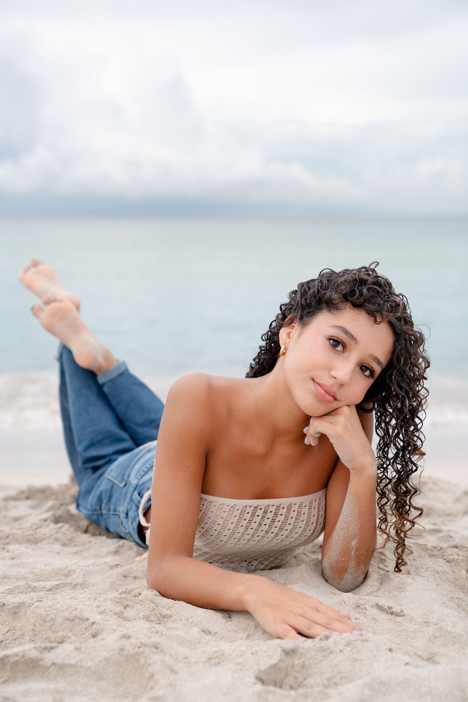 Young woman with curly hair lying on sandy beach in jeans and strapless top, ocean in background.