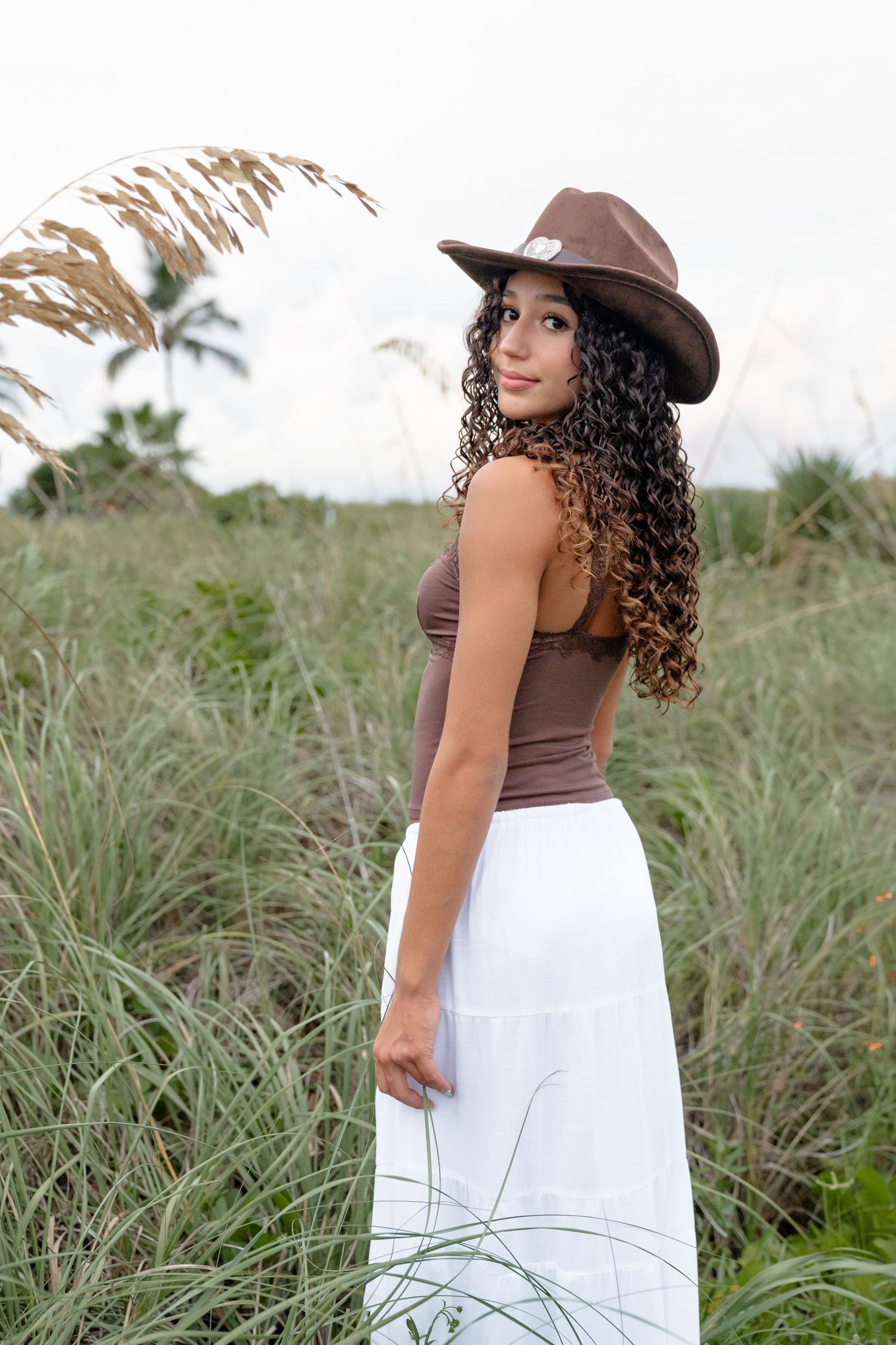 Young woman with curly hair wearing a brown cowboy hat, brown top, and white maxi skirt standing in tall coastal grass.