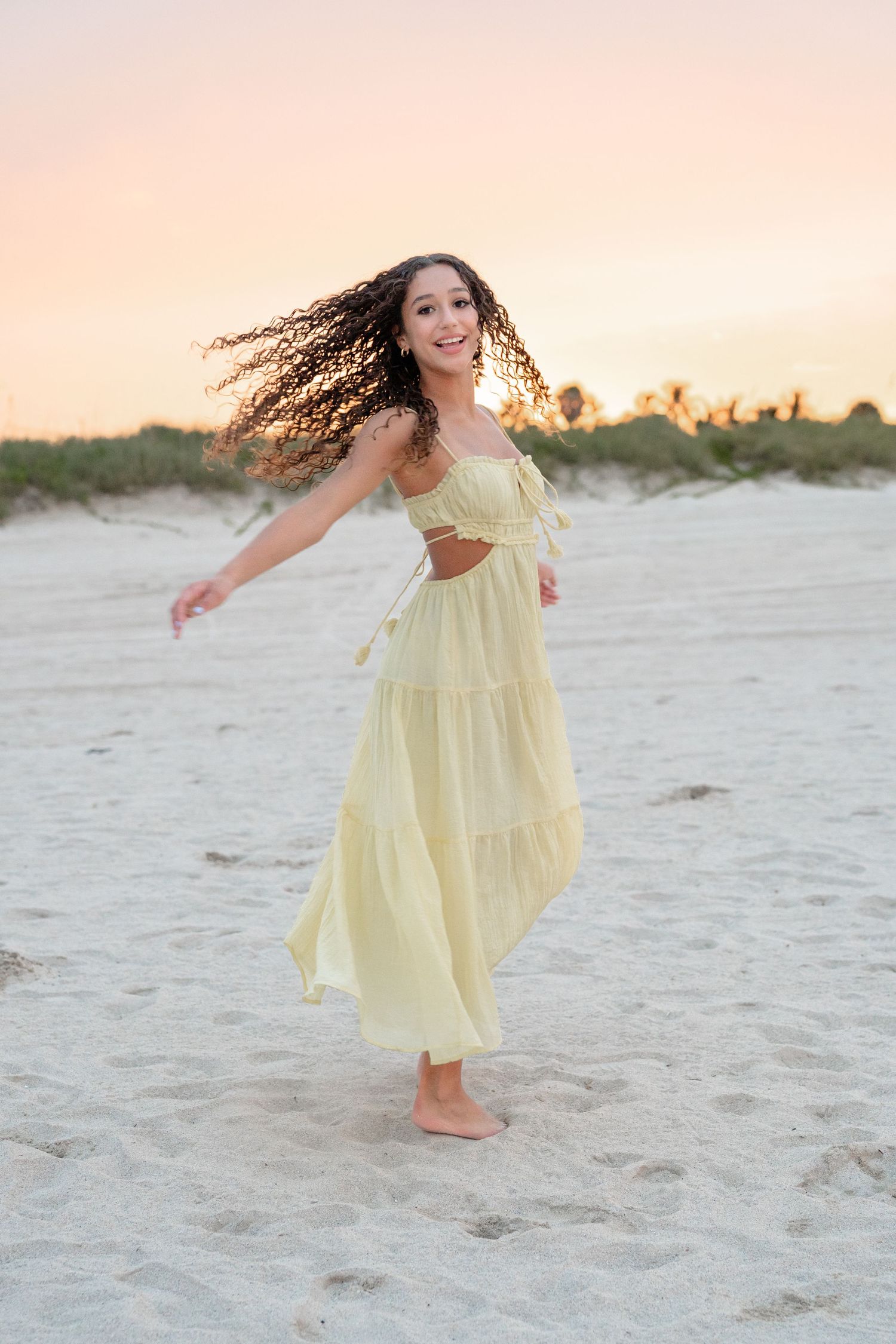 Young woman in yellow dress dancing barefoot on sandy beach at sunset with hair flowing.