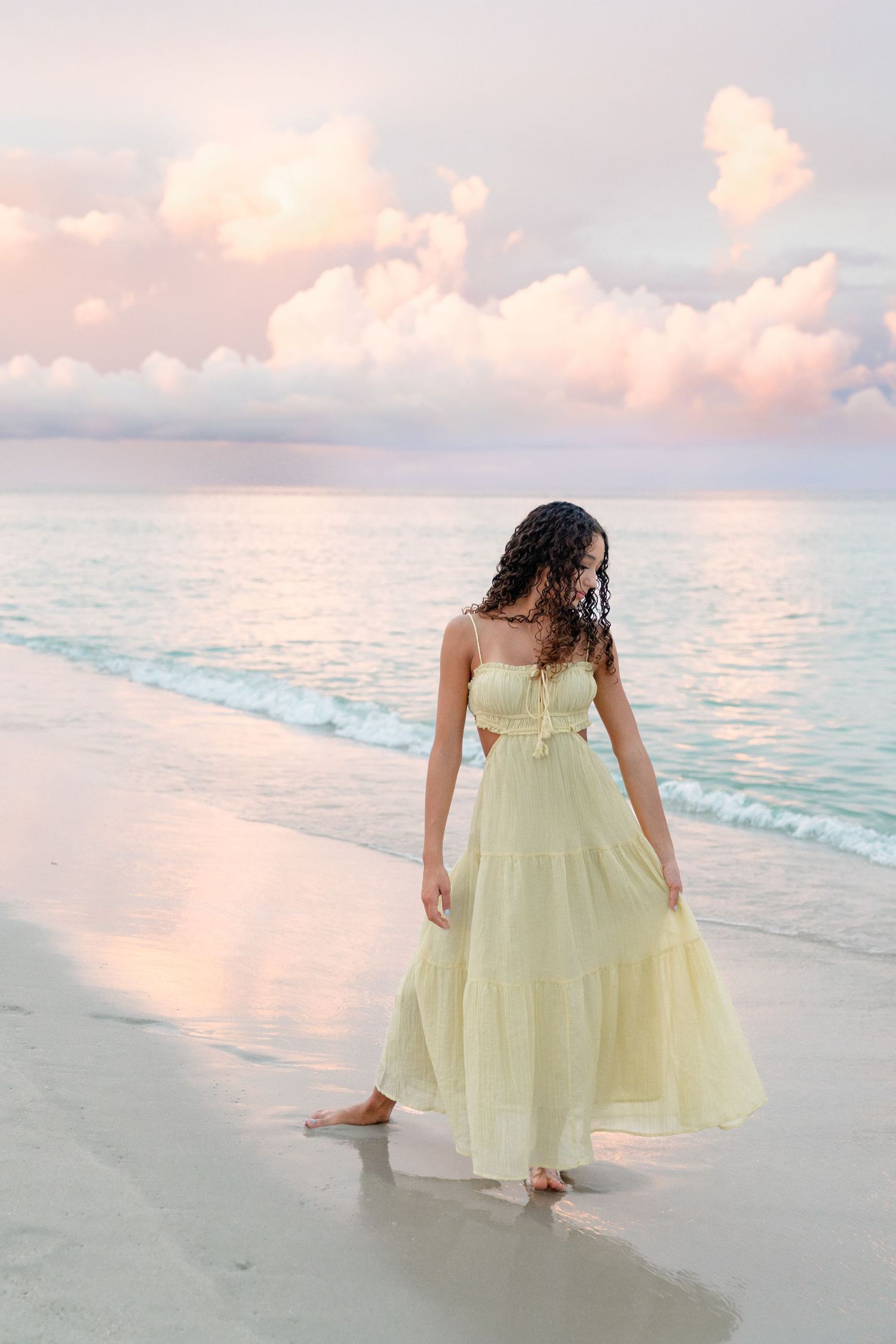 Woman in yellow maxi dress walking barefoot on beach at sunset with pink clouds reflecting on calm ocean water.