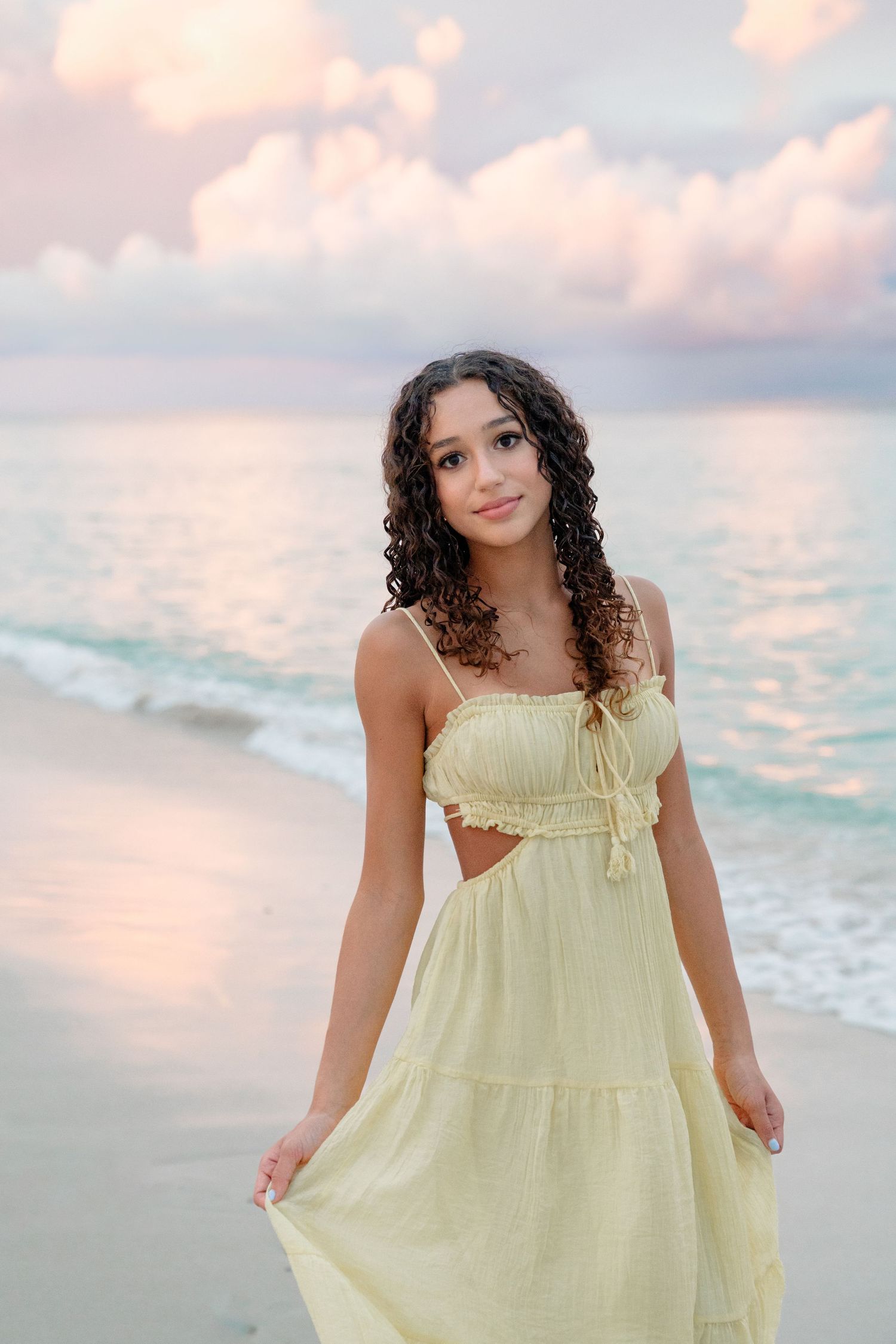 Young woman in yellow sundress poses on a tropical beach at sunset with turquoise waves and pink clouds.