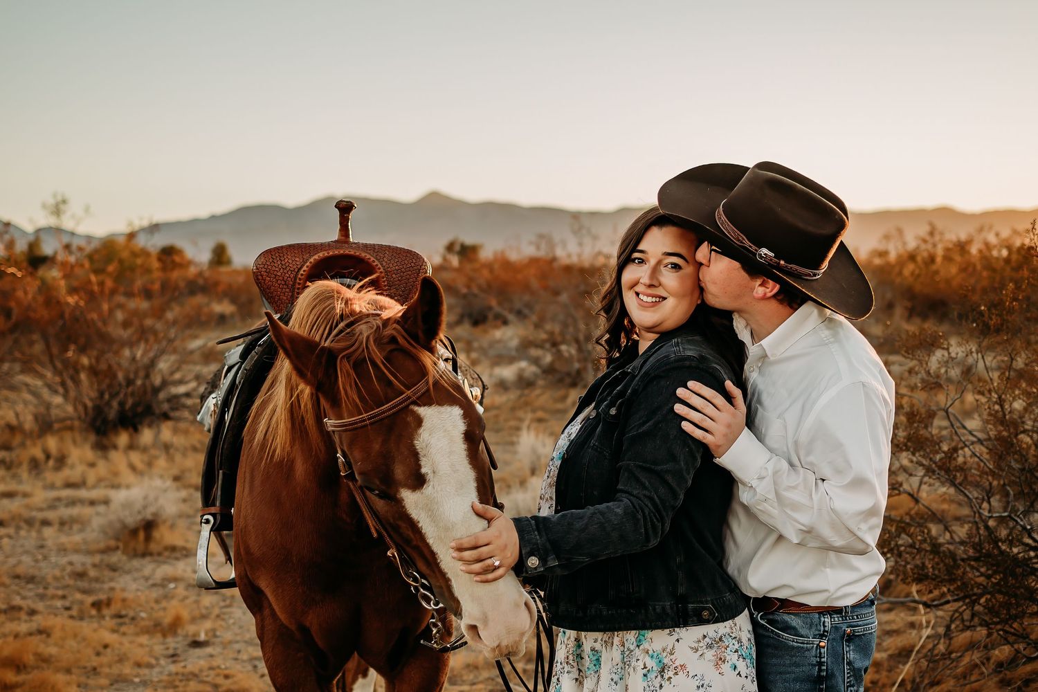 Couple in cowboy hats sharing a kiss beside a chestnut horse in a desert landscape at golden hour.