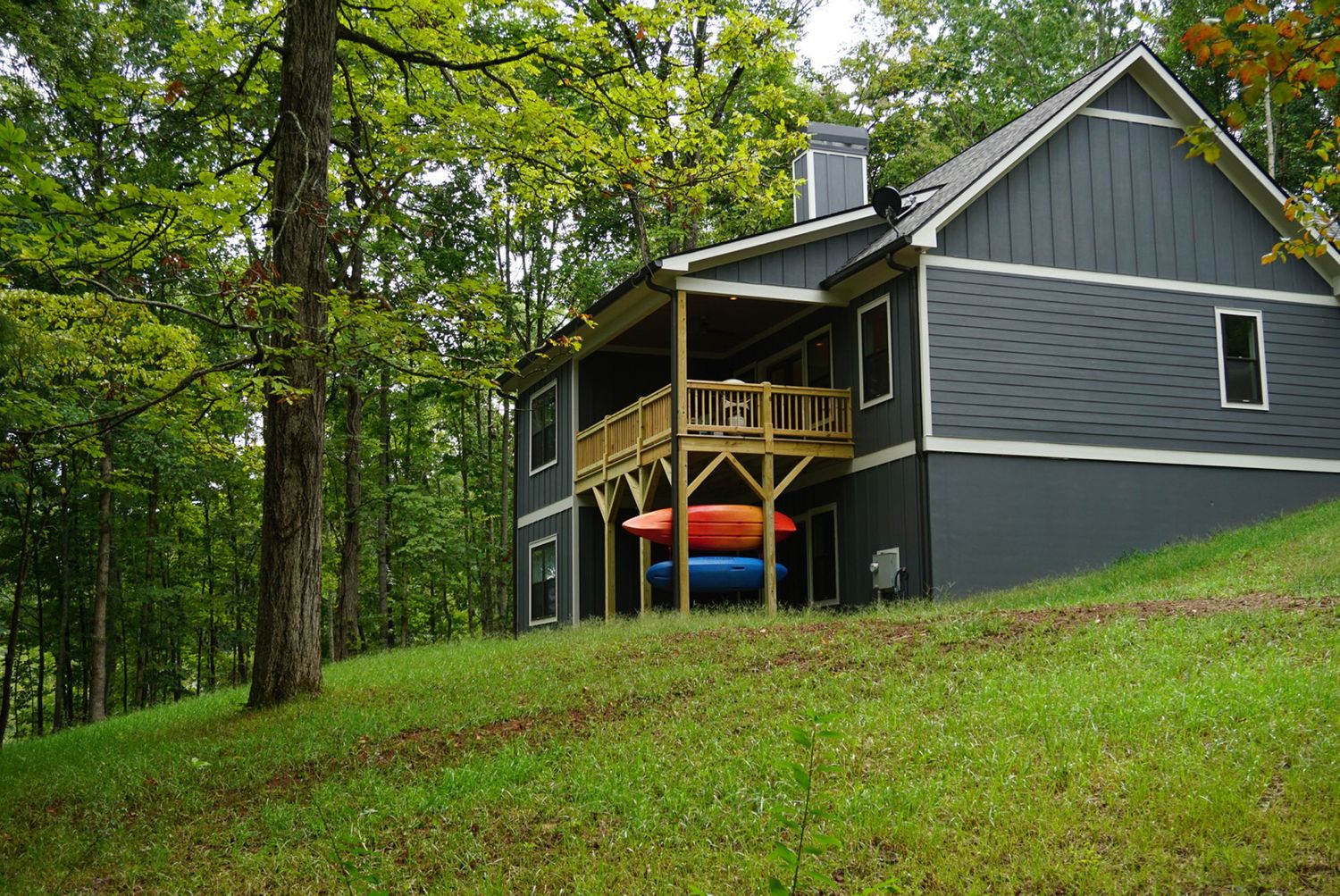 A grey home with white trim and two kayaks sits on a grassy hill surrounded by trees on Lake Chatuge in Hiawassee, GA .
