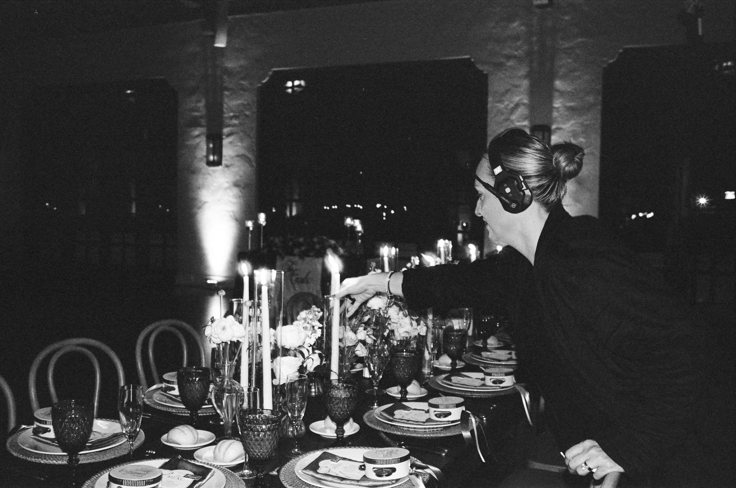 Black and white photo of a woman arranging flowers at an elegant candlelit dinner table set for a formal event.