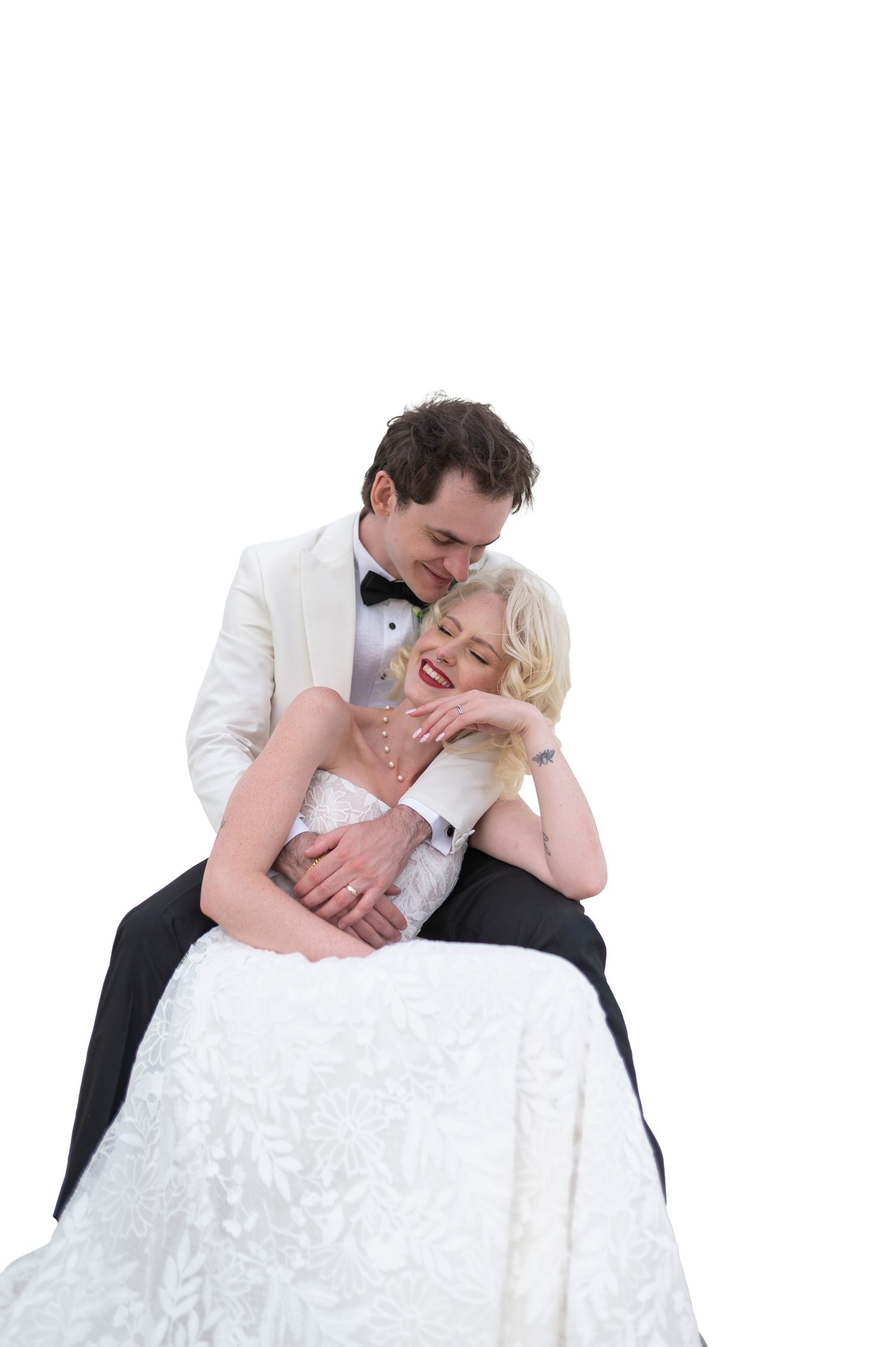 A joyful bride in a white gown sits on her groom's lap as he embraces her tenderly in a wedding portrait.