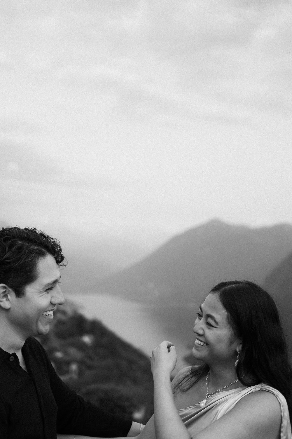 Black and white photo of a smiling couple laughing together with scenic mountain lake backdrop.