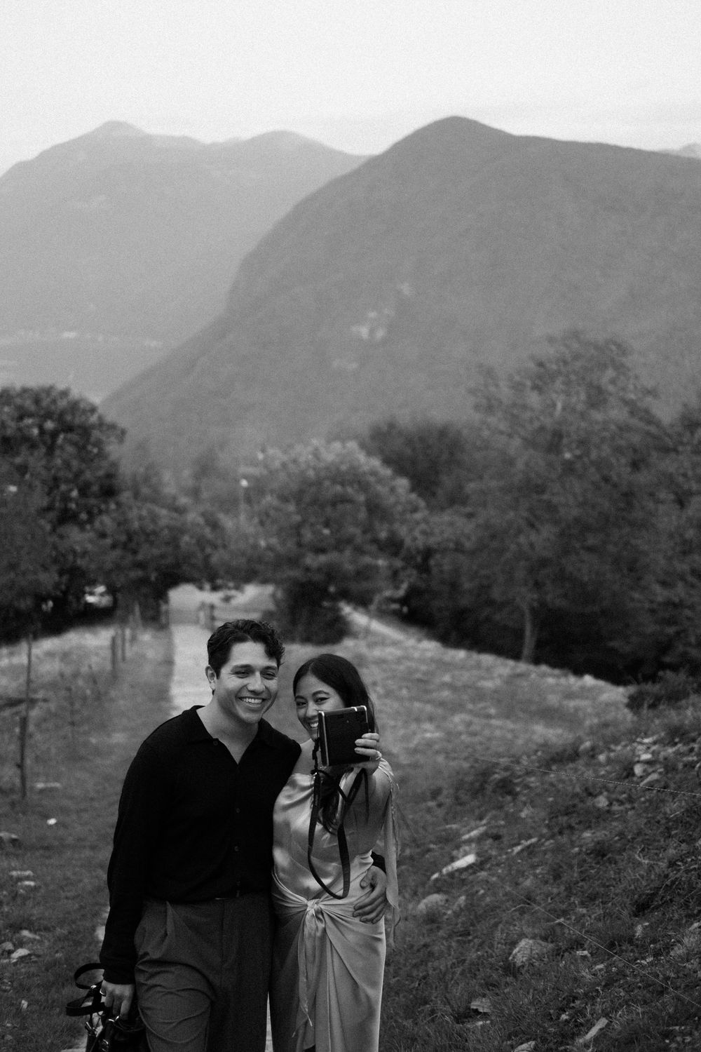Black and white photo of a smiling couple posing together with a camera in front of a scenic mountain landscape.