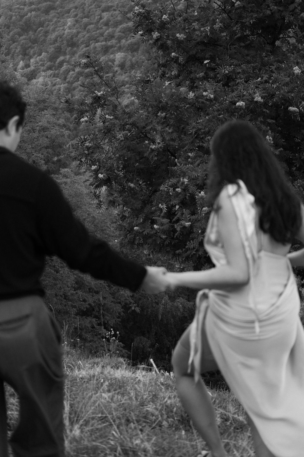 Black and white photo of a couple holding hands walking through a lush green hillside meadow.
