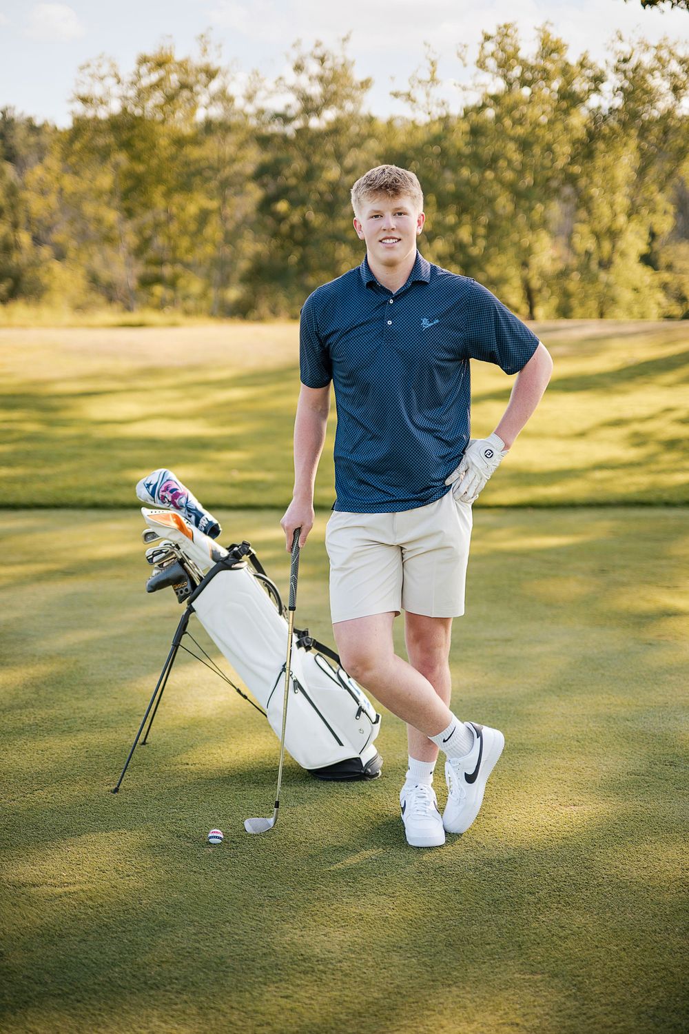 Young male golfer in navy polo and khaki shorts posing with golf bag on a sunny green course.