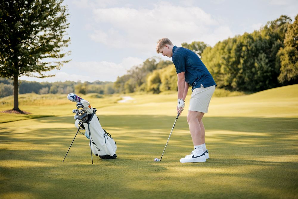 Young man in blue shirt and khaki shorts playing golf on a lush green course with a golf bag nearby.