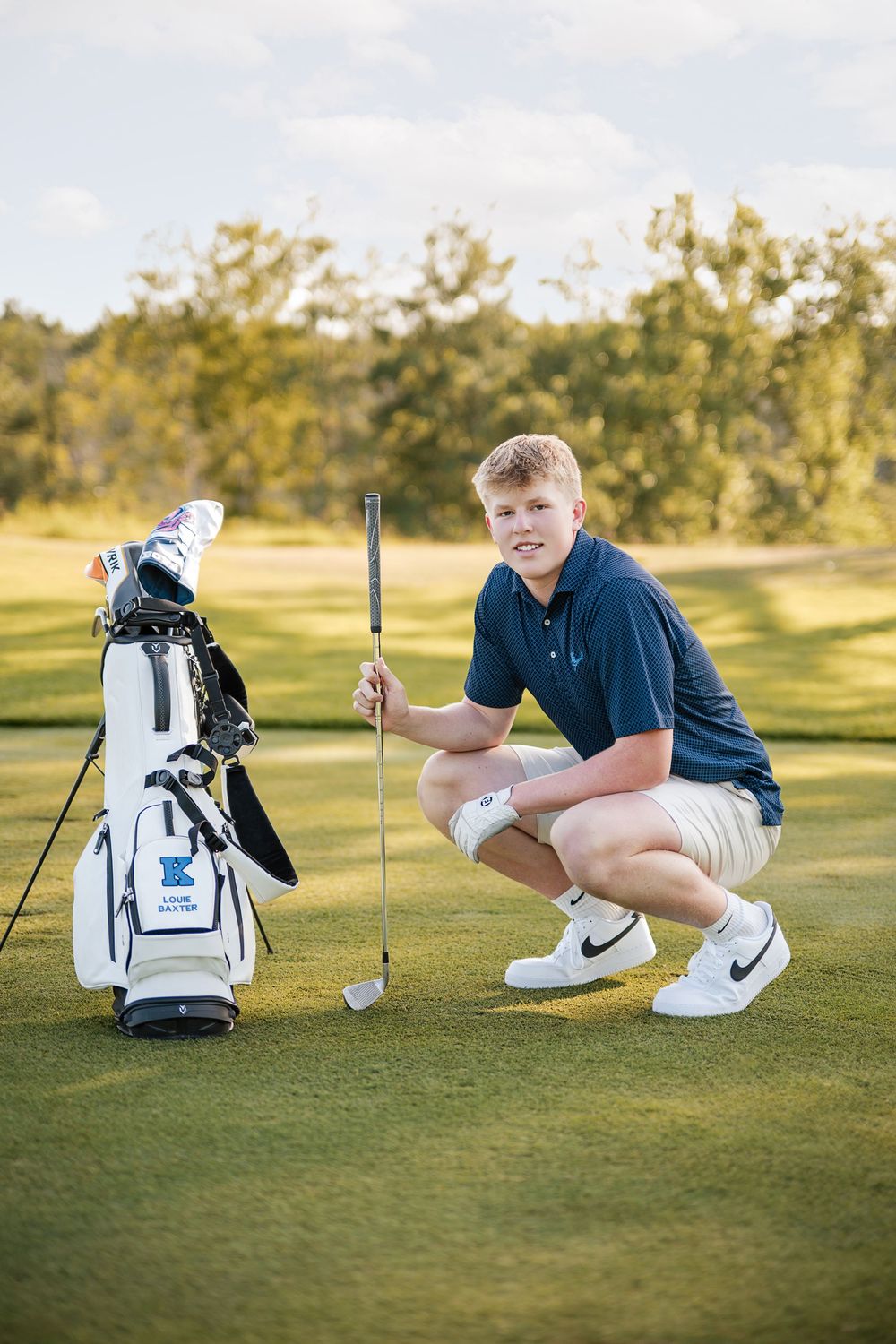 Young male golfer crouching on green beside golf bag, holding club, wearing navy polo and Nike shoes.