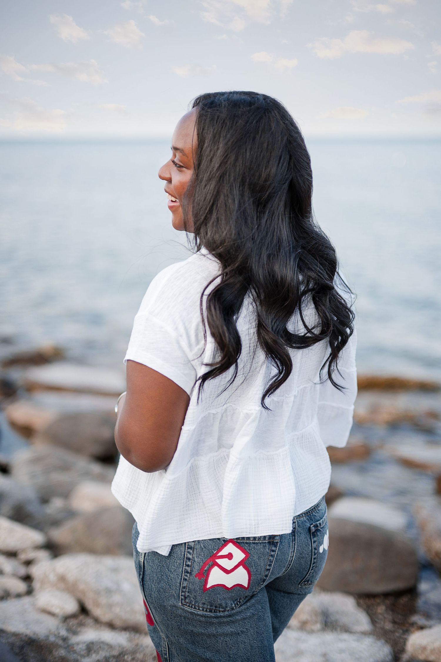 Young Black woman with long wavy hair in white top and jeans smiling by rocky shoreline.