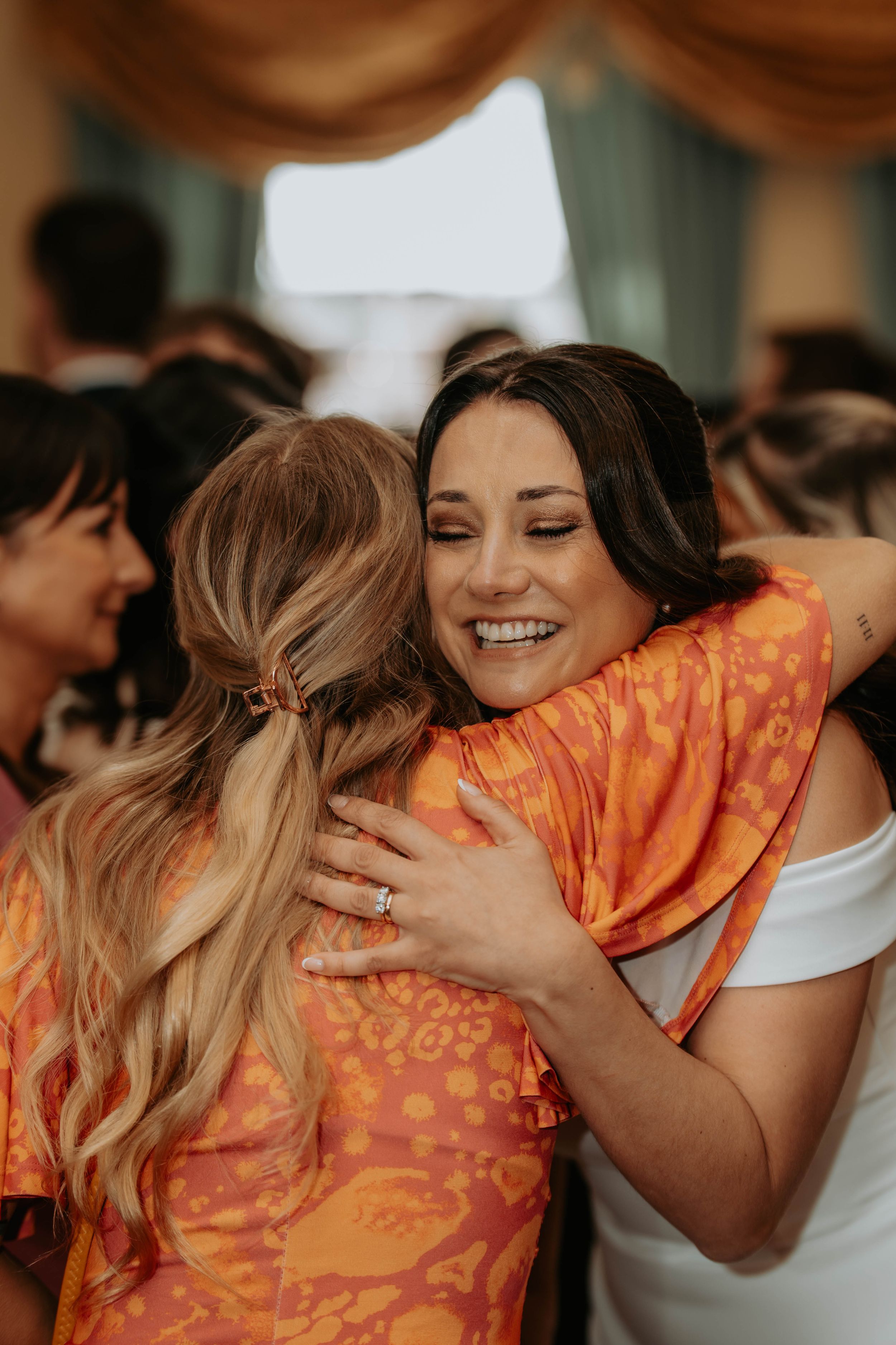 Two women share a warm embrace at a celebration, one wearing an orange floral dress, smiling joyfully.