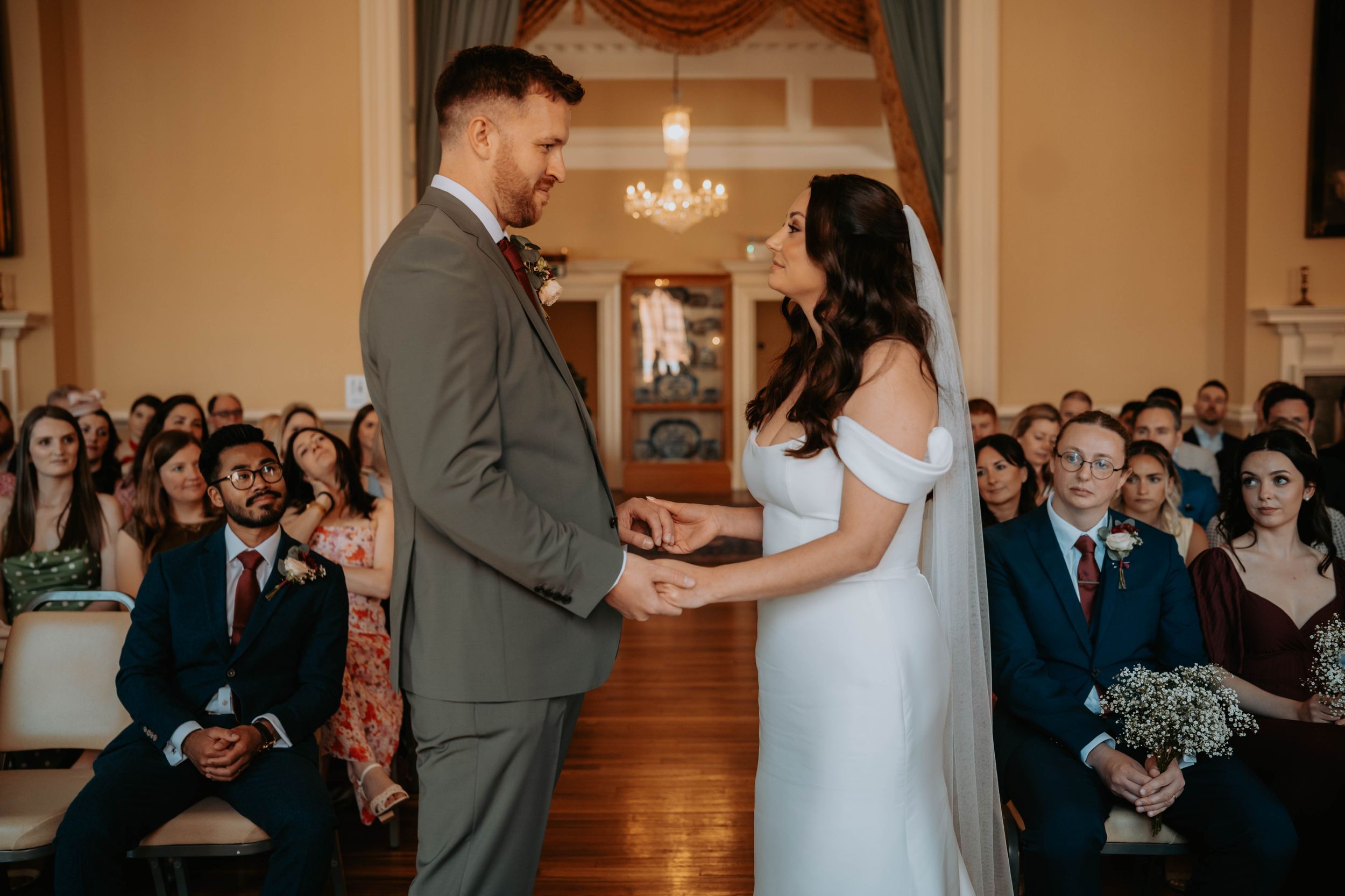 Bride and groom exchange vows at elegant indoor wedding ceremony with guests seated in background.