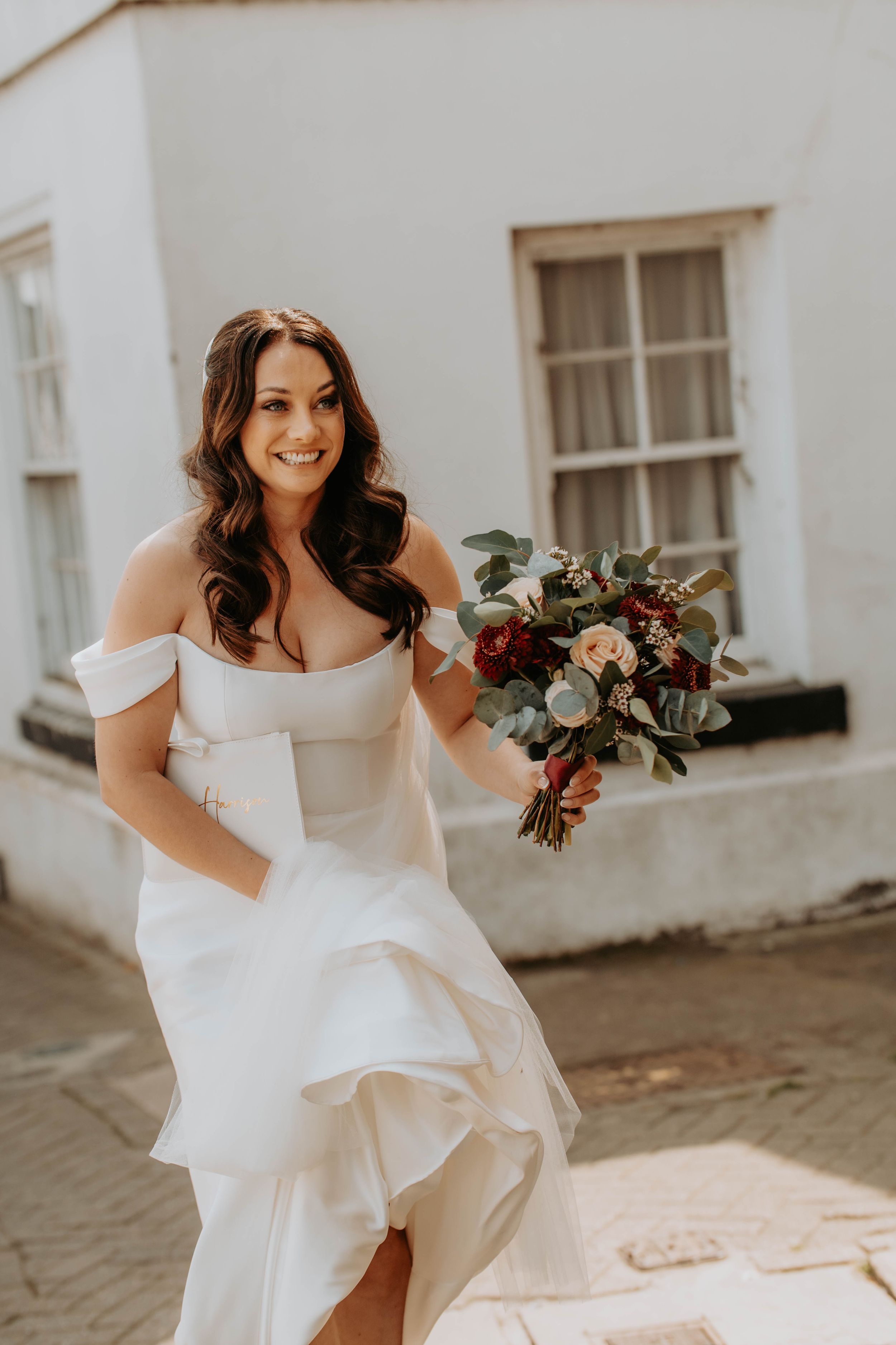 Smiling bride in off-shoulder white gown holding a burgundy and blush bouquet, posing outdoors near a white building.