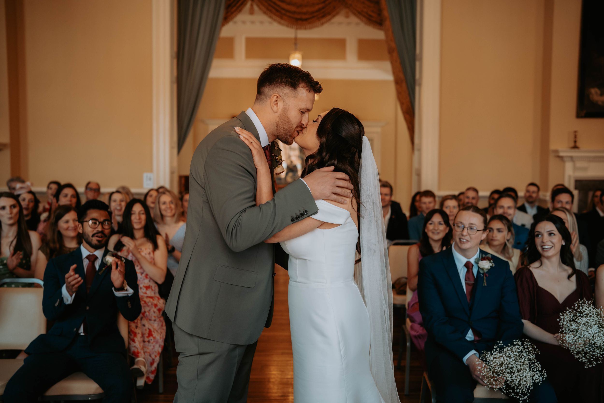 Bride and groom share first kiss at wedding ceremony altar with guests applauding in background.