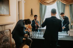 Men in suits preparing champagne glasses on a table by elegant curtained windows at a formal event.