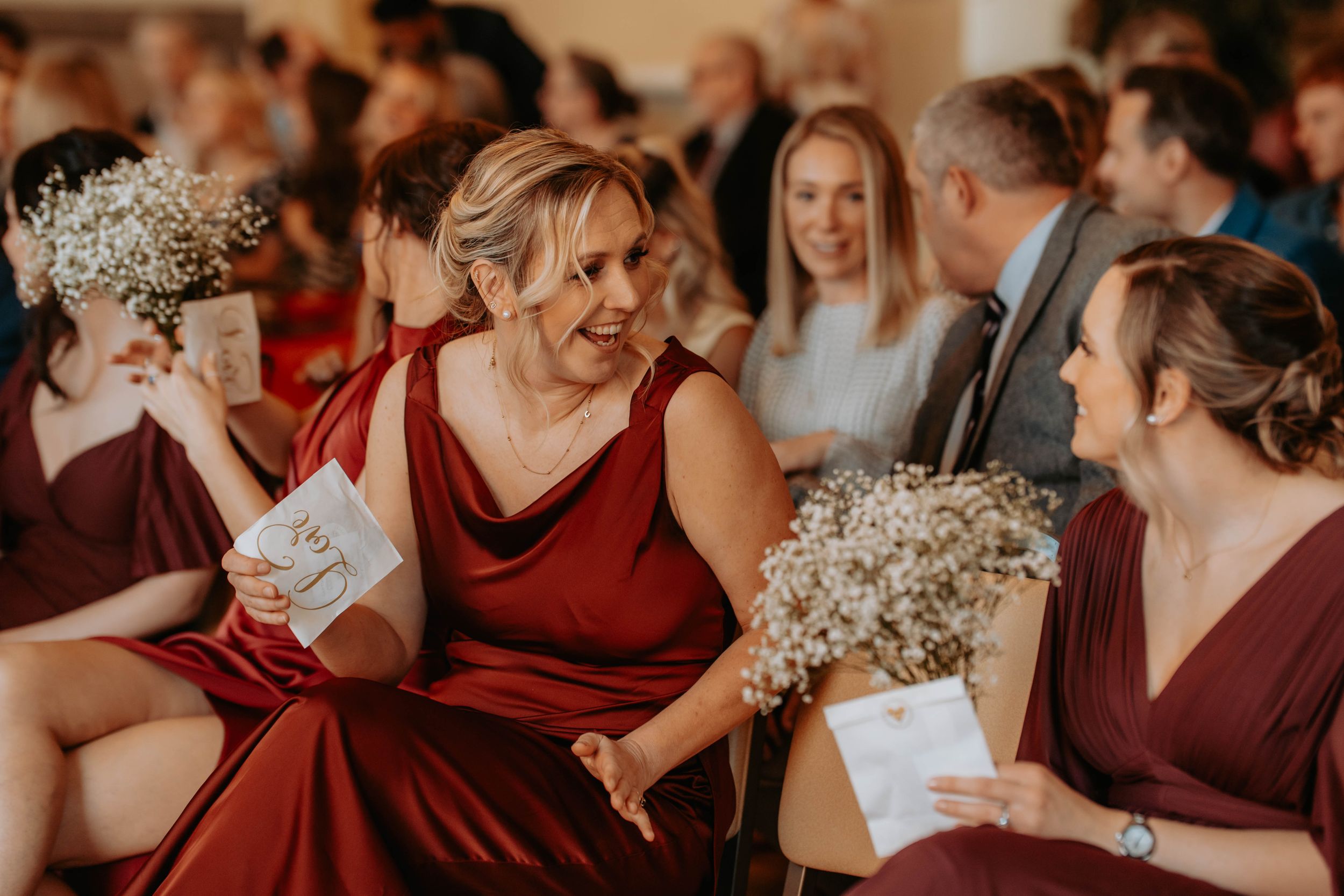 Bridesmaids in burgundy dresses holding baby's breath bouquets and programs, laughing during a wedding ceremony.