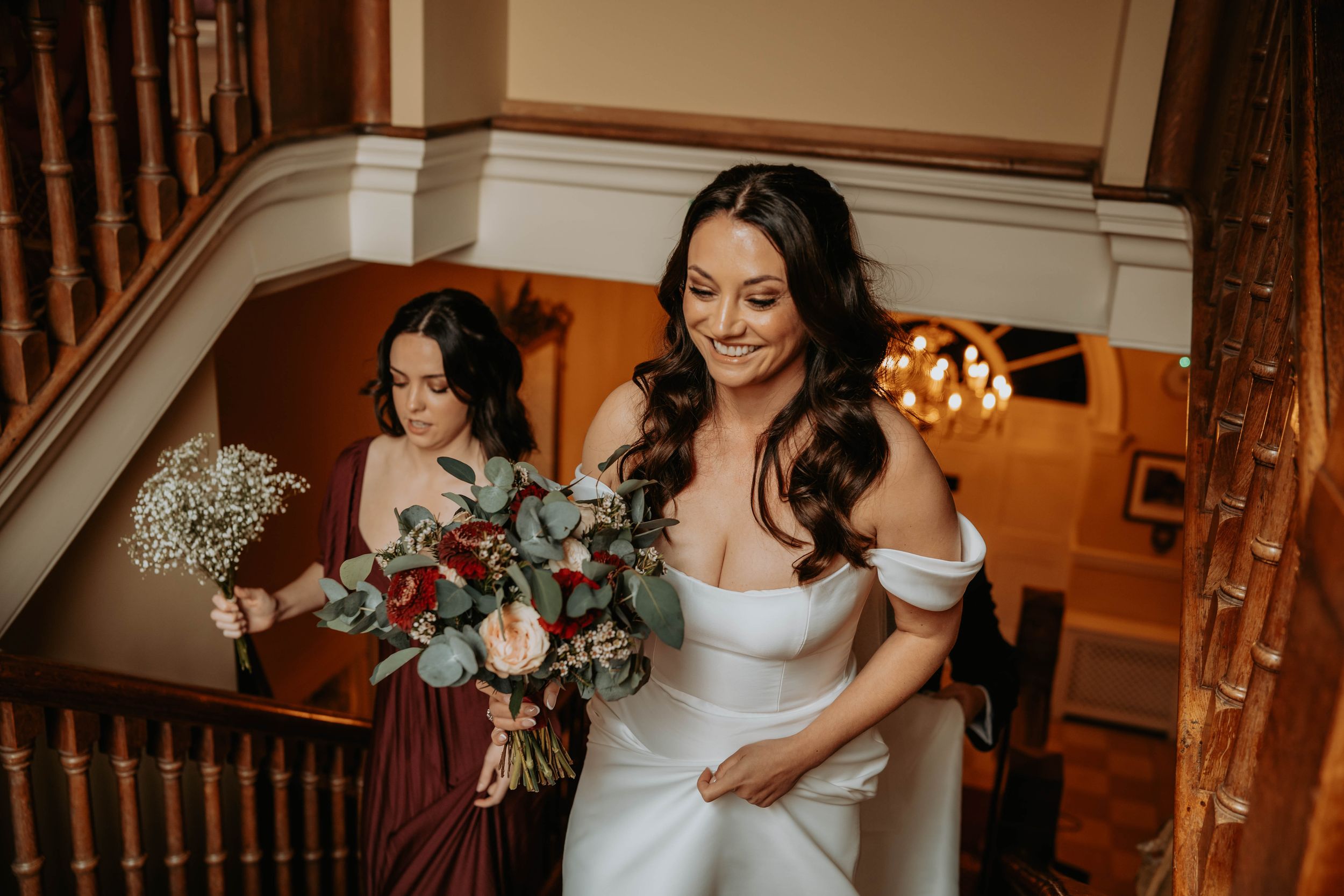 Bride in white off-shoulder gown descends staircase with bridesmaid in burgundy dress holding bouquets at wedding venue.