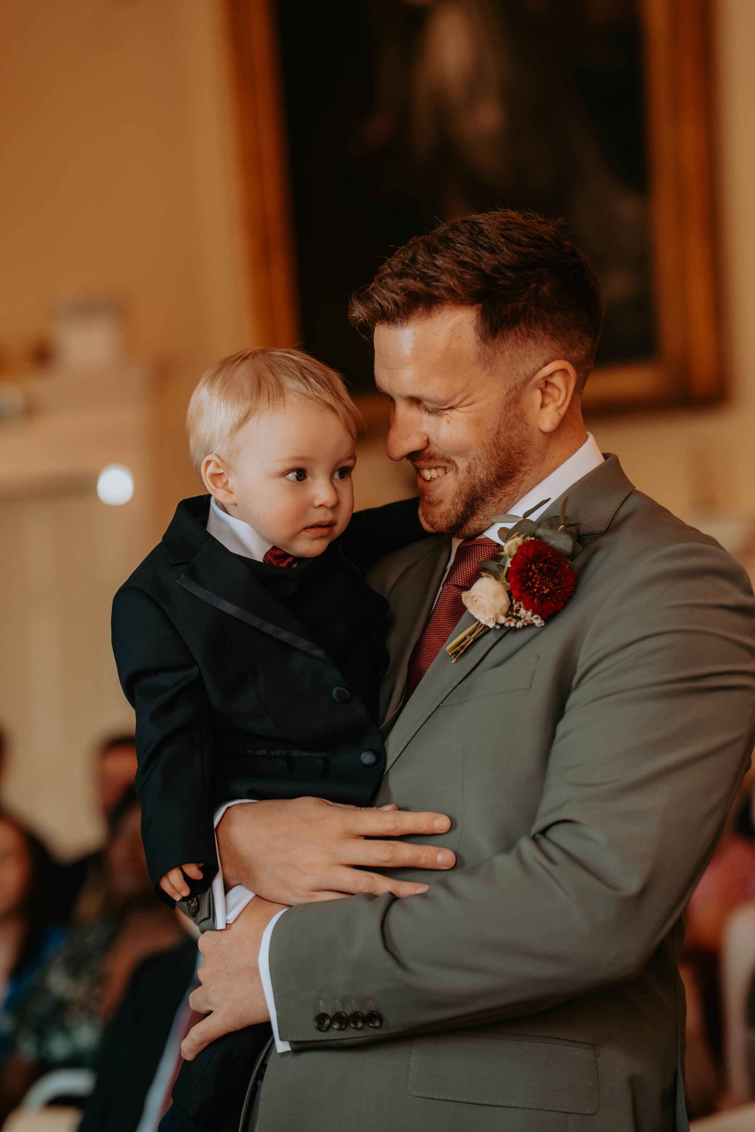 Smiling groom in gray suit holds a toddler boy in a black tuxedo during a wedding ceremony indoors.