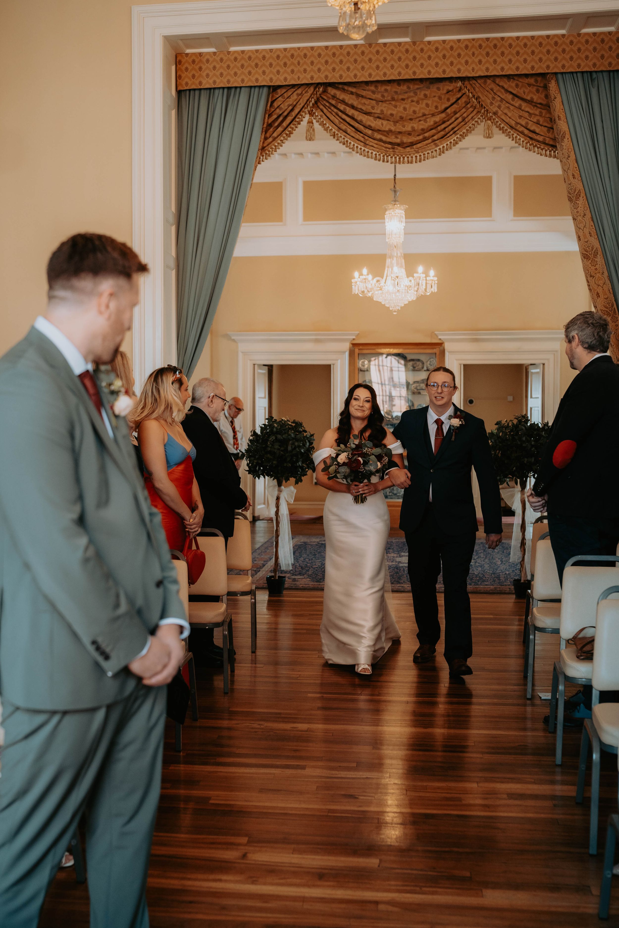 Bride walks down the aisle with escort in elegant venue, chandelier overhead, groom awaiting at ceremony.