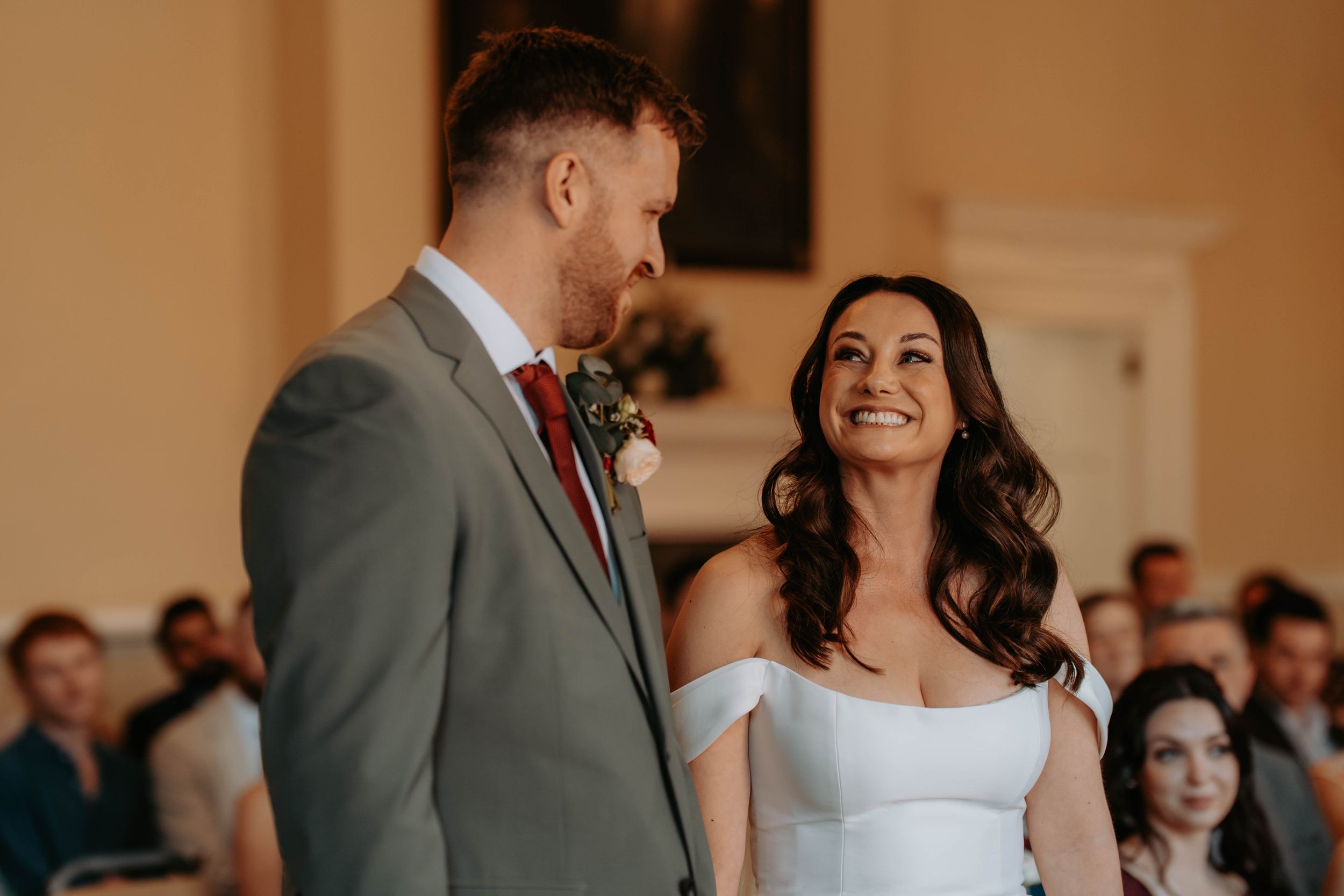 A bride in a white off-shoulder gown smiles joyfully at her groom in a gray suit during a wedding ceremony.