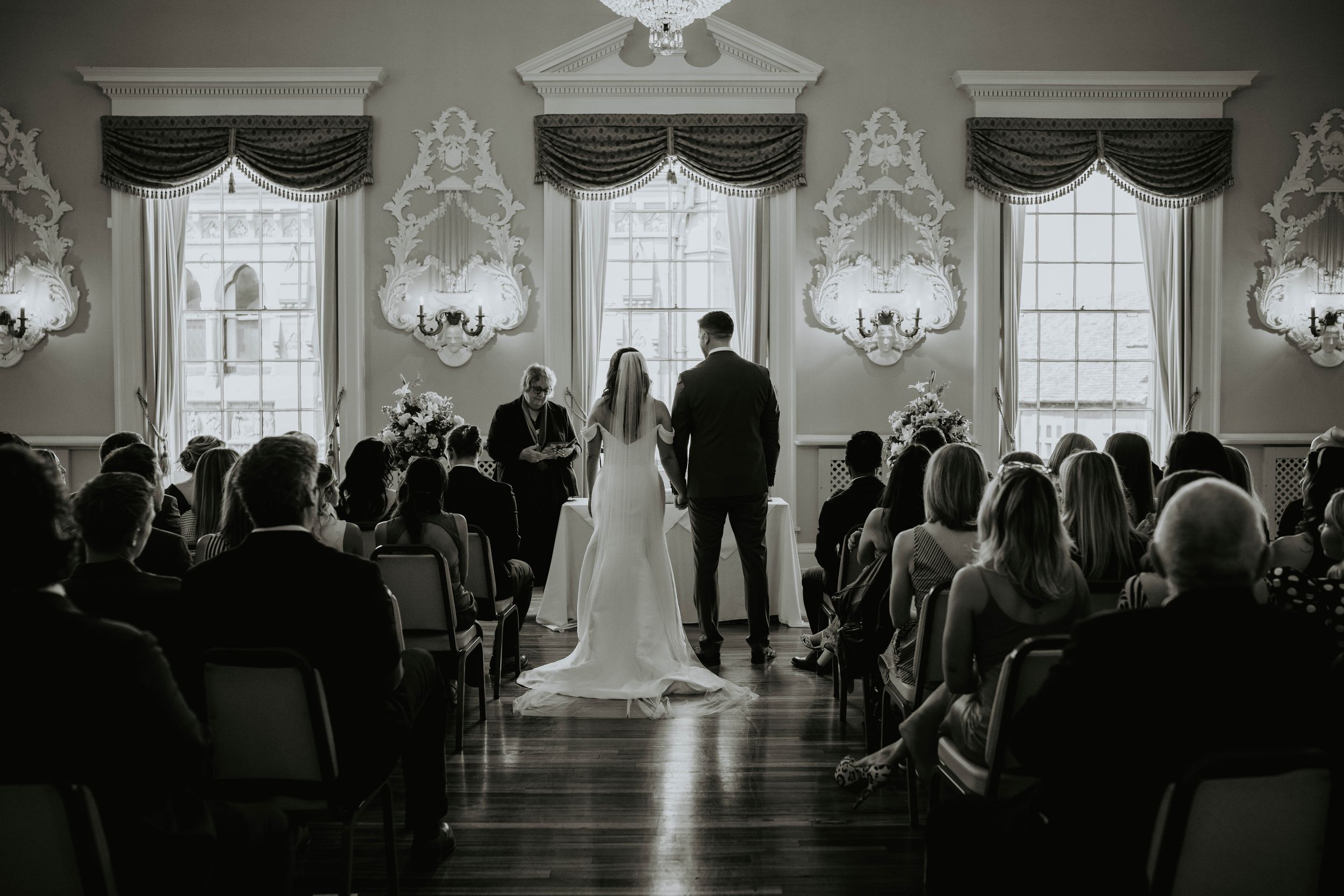 Black and white wedding ceremony photo of bride and groom at altar with seated guests in elegant ballroom.