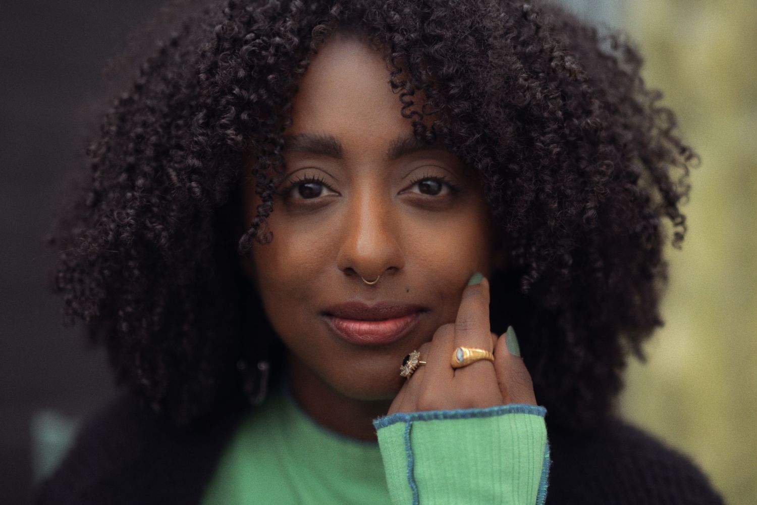 Young Black woman with natural curly hair wearing a green turtleneck and gold ring, posing outdoors.