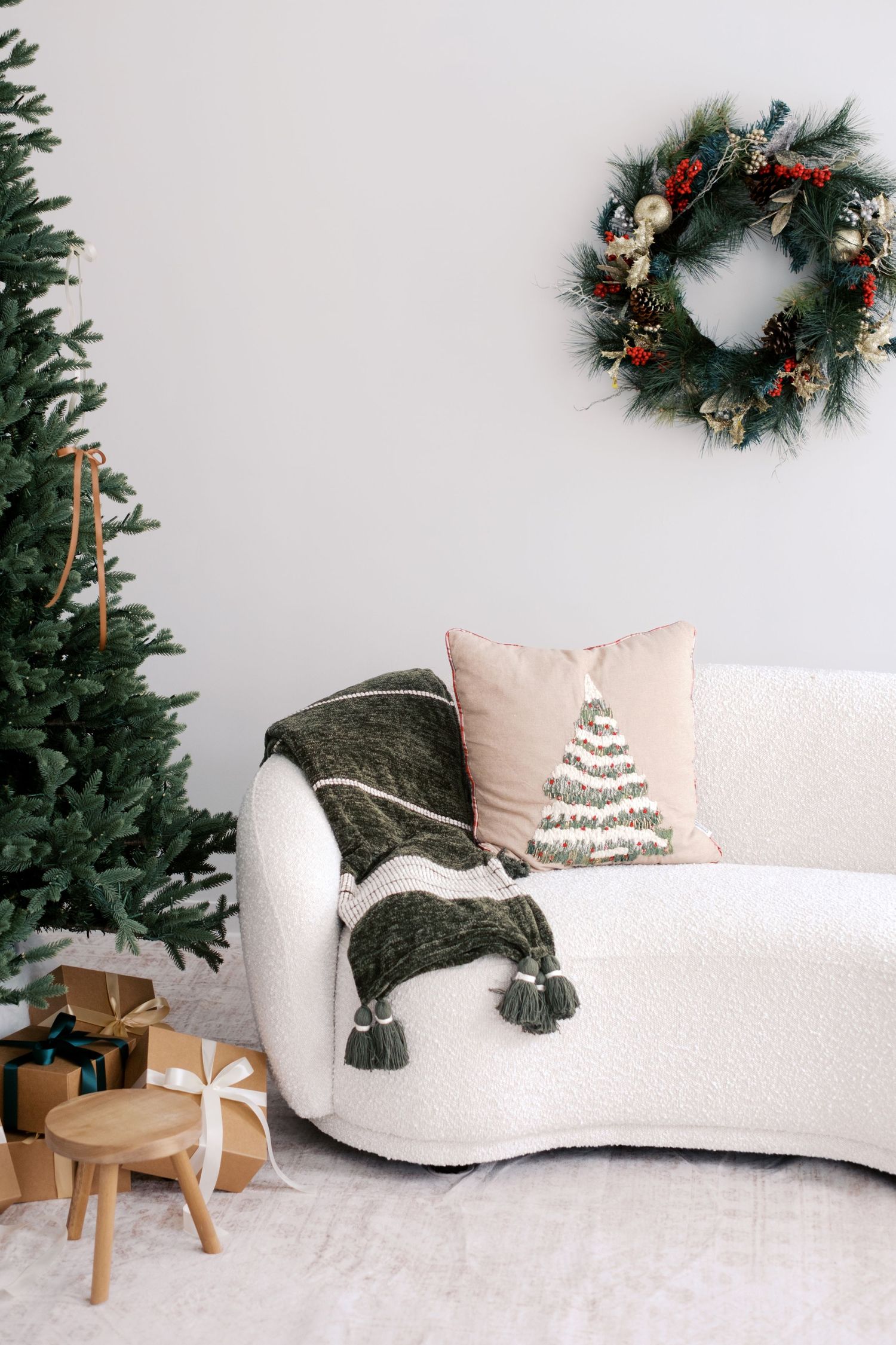 Cozy Christmas living room with white boucle sofa, green throw, festive wreath, decorated tree, and wrapped gifts.