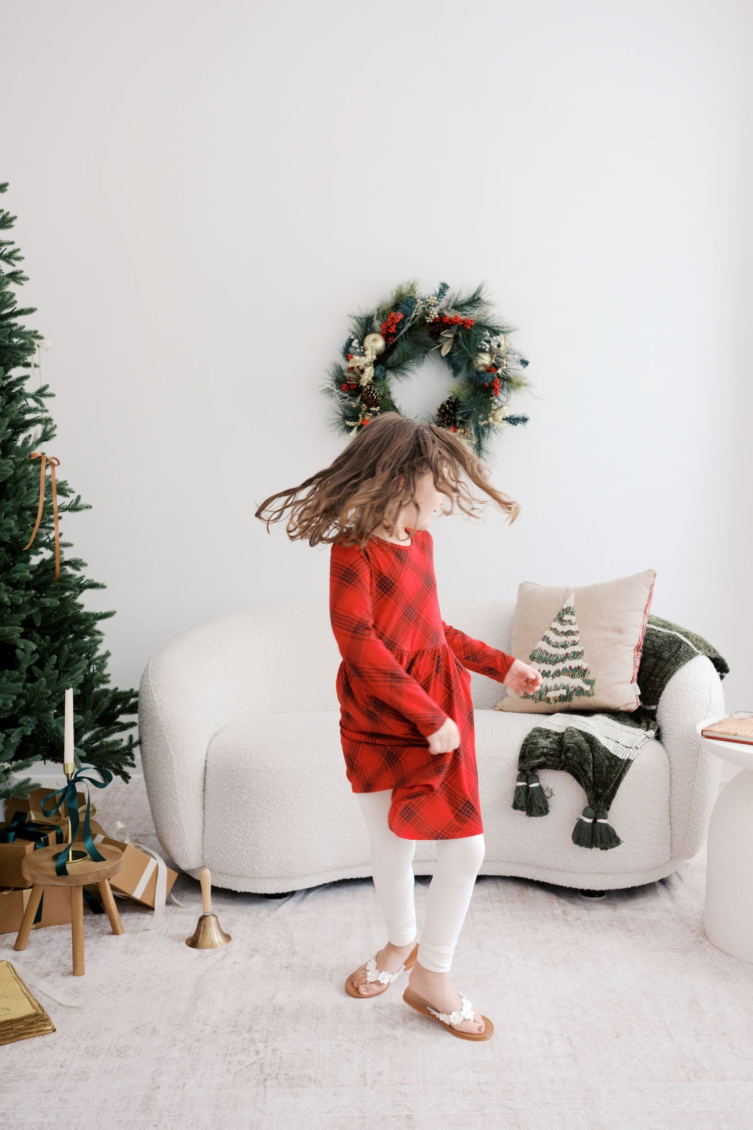 A young girl in a red dress twirls joyfully near a Christmas tree and festive wreath in a decorated living room.