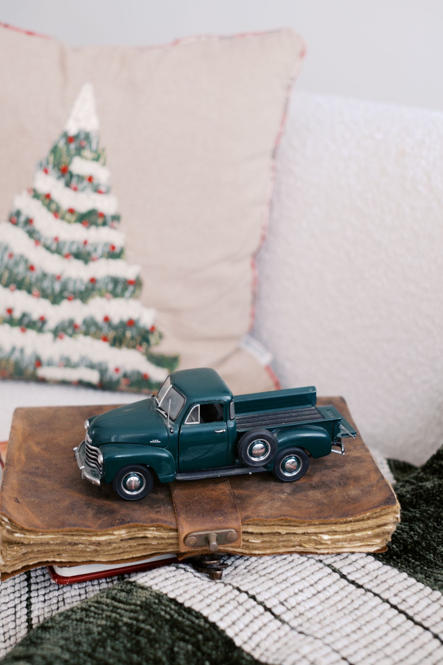 A vintage green toy pickup truck sits atop an old leather book with Christmas pillows in the background.