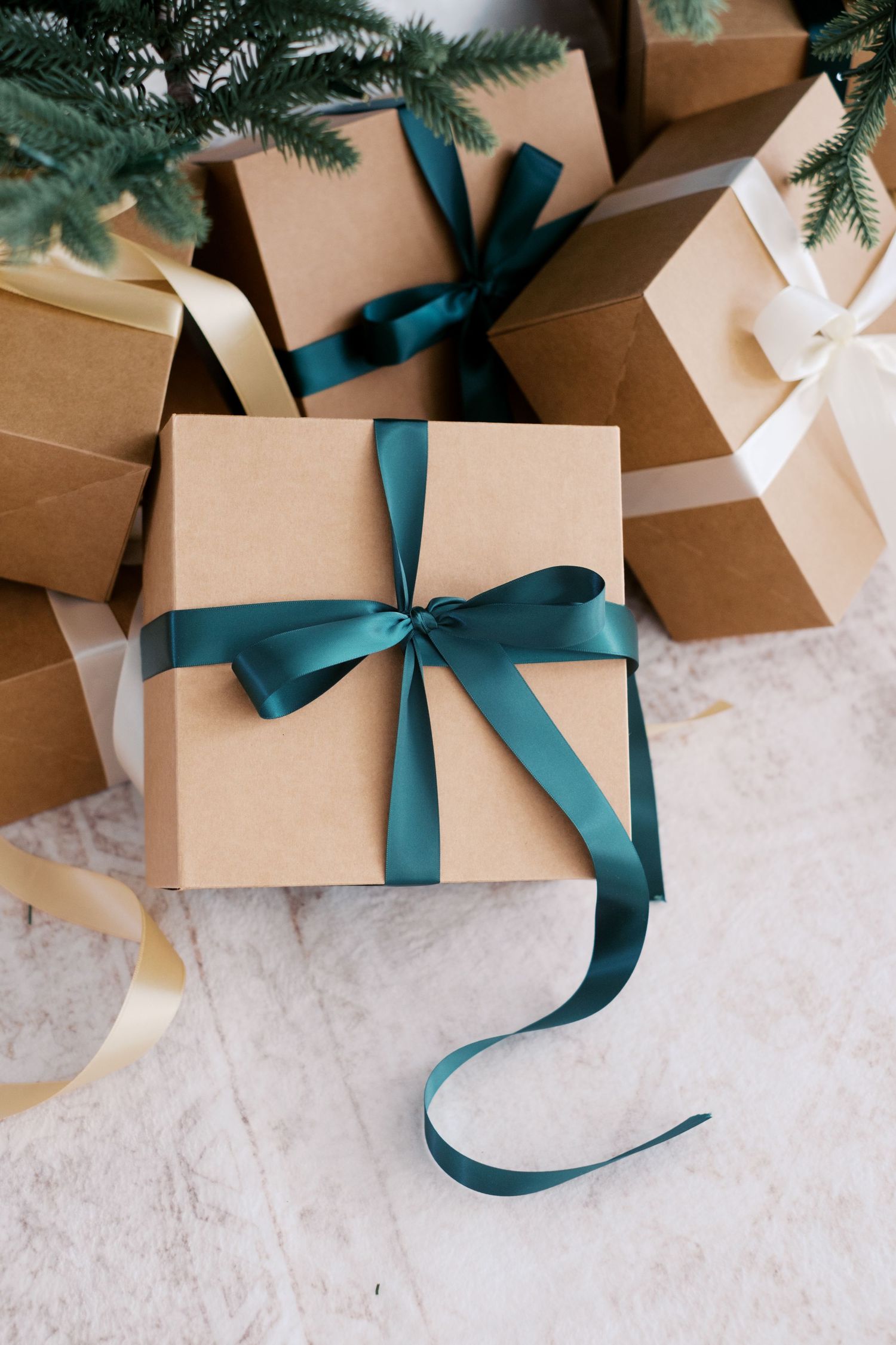 Kraft paper gift boxes with teal and gold satin ribbons arranged under a Christmas tree on a white surface.