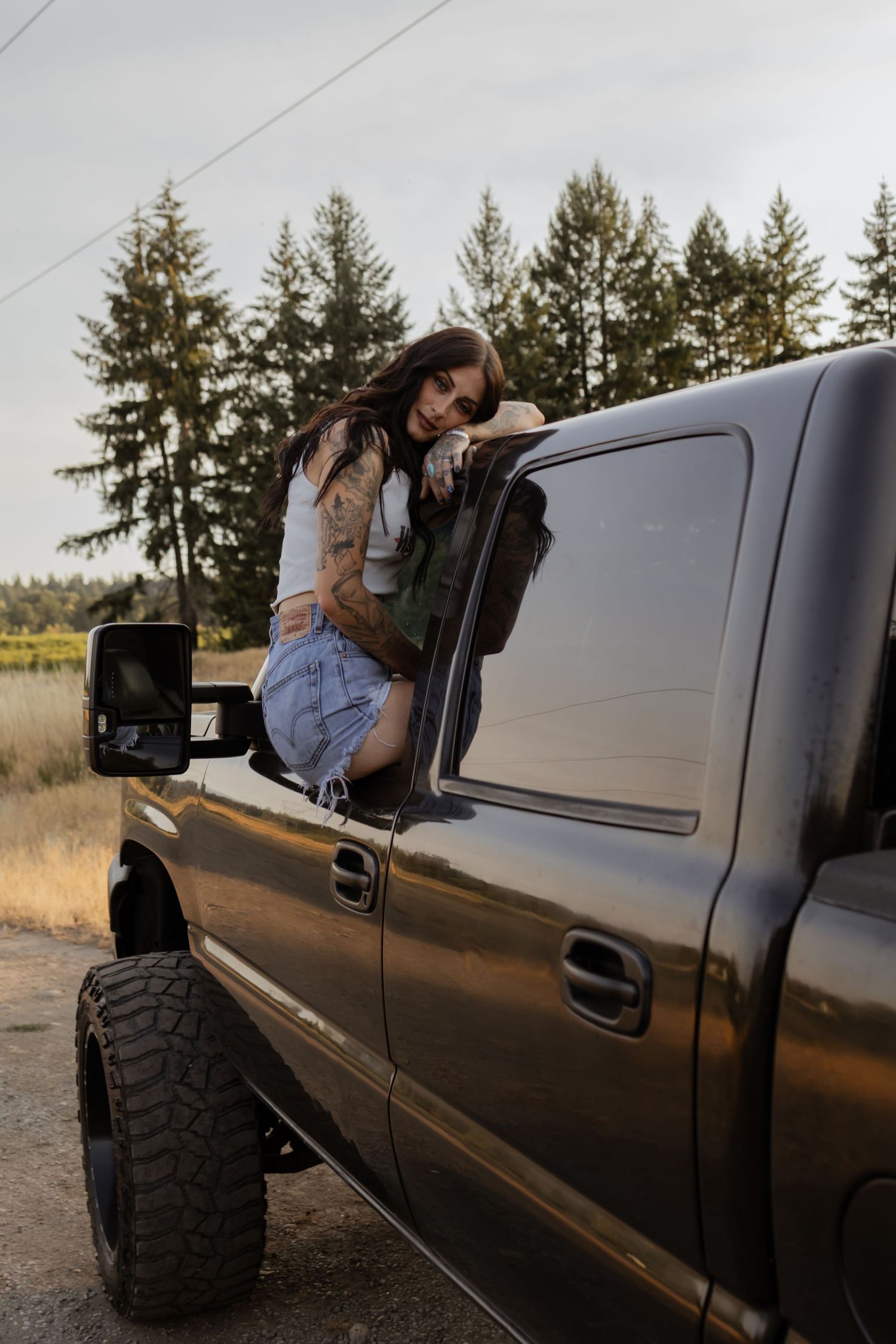 A woman in denim shorts leans out of a lifted dark pickup truck surrounded by tall evergreen trees.