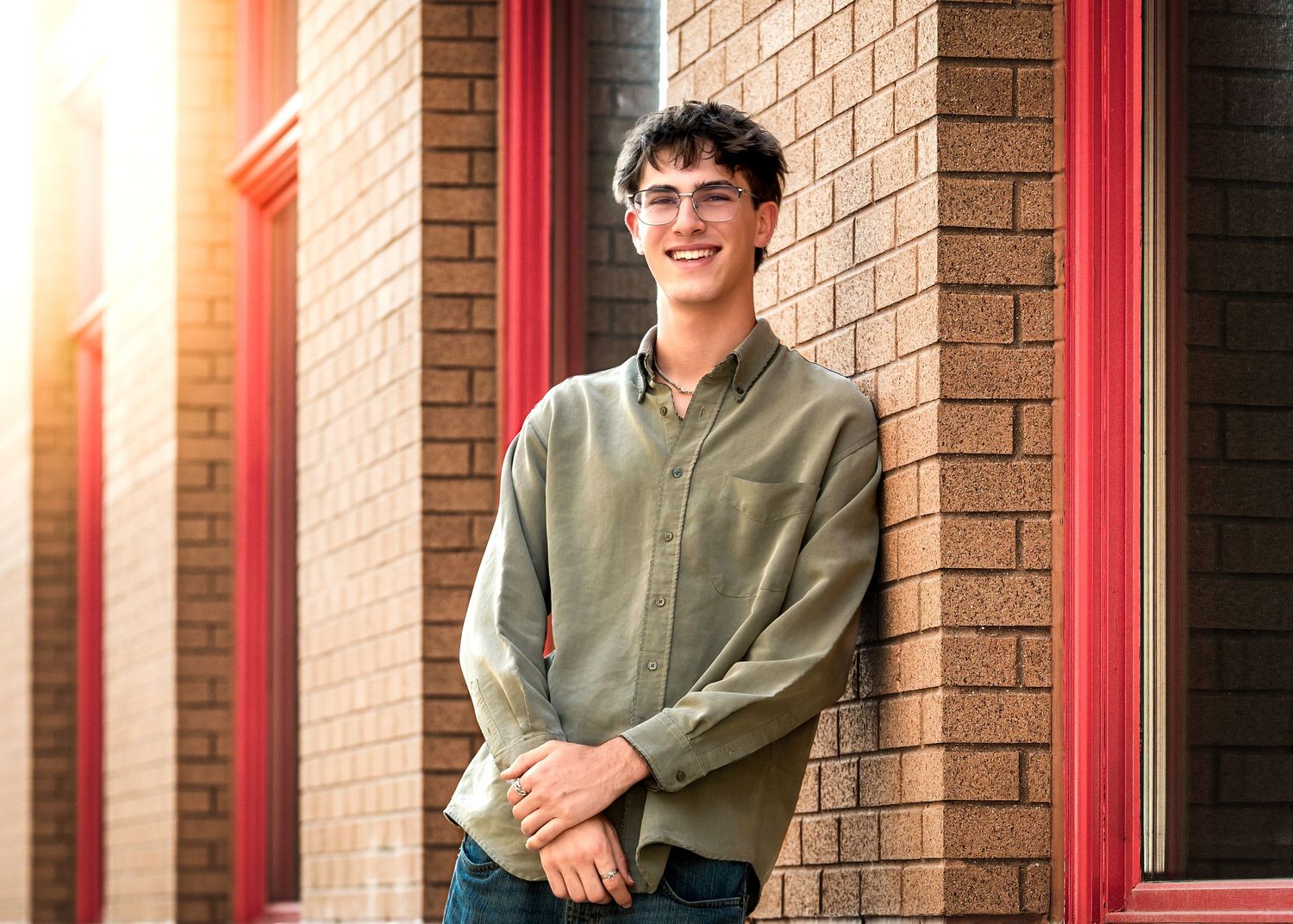 Young man in olive shirt and glasses smiling against a brick wall in warm sunlight.