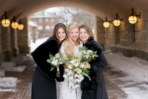Bride and two bridesmaids in black fur wraps holding white bouquets under a snowy archway.