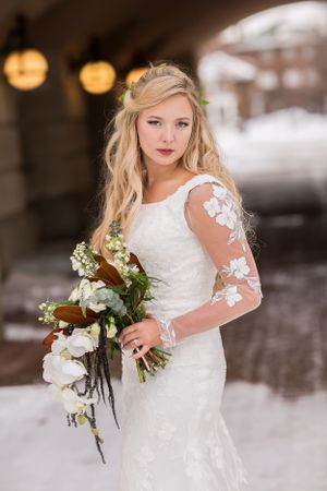 Bride in white lace long-sleeve gown holding floral bouquet outdoors in snowy winter setting.