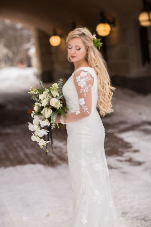 Bride in lace wedding gown holding white floral bouquet, posing outdoors in snowy winter setting.