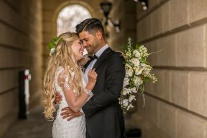 Bride and groom embrace tenderly in an elegant archway, she holds a white floral bouquet, both dressed in wedding attire.