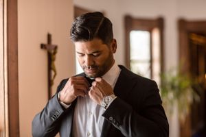 Groom in black suit adjusting bow tie before wedding ceremony with crucifix on wall in background.