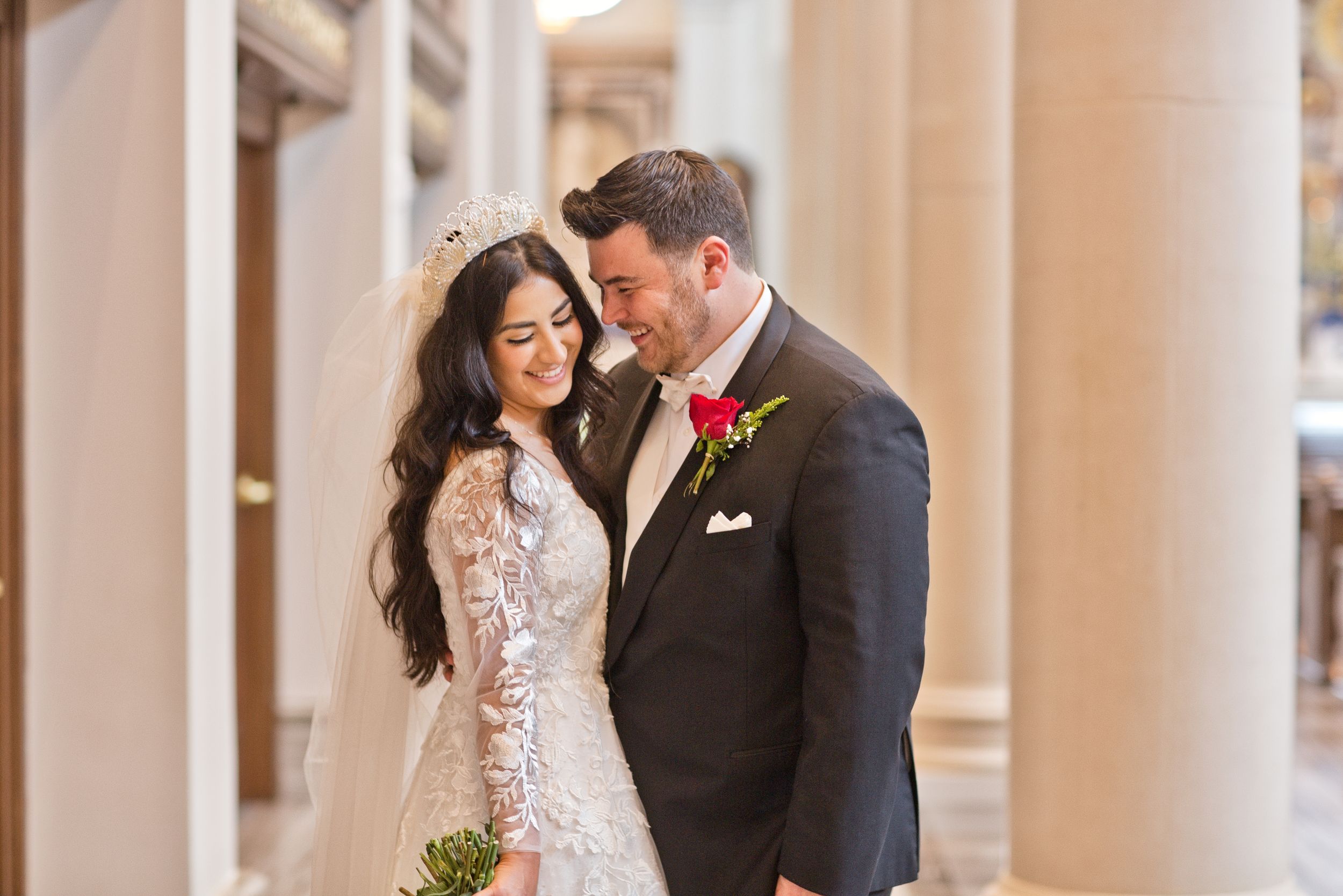 Elegant bride in lace gown and groom in dark suit share a tender moment among grand columns.